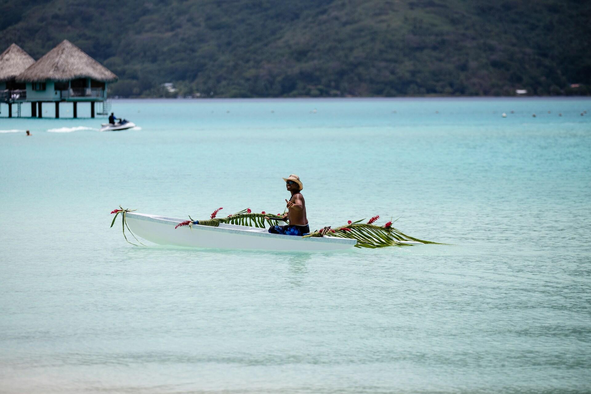 Una persona con un sombrero de paja rema en una canoa decorada sobre aguas tranquilas de color turquesa, cerca de cabañas tropicales sobre pilotes y de una costa montañosa y frondosa.
