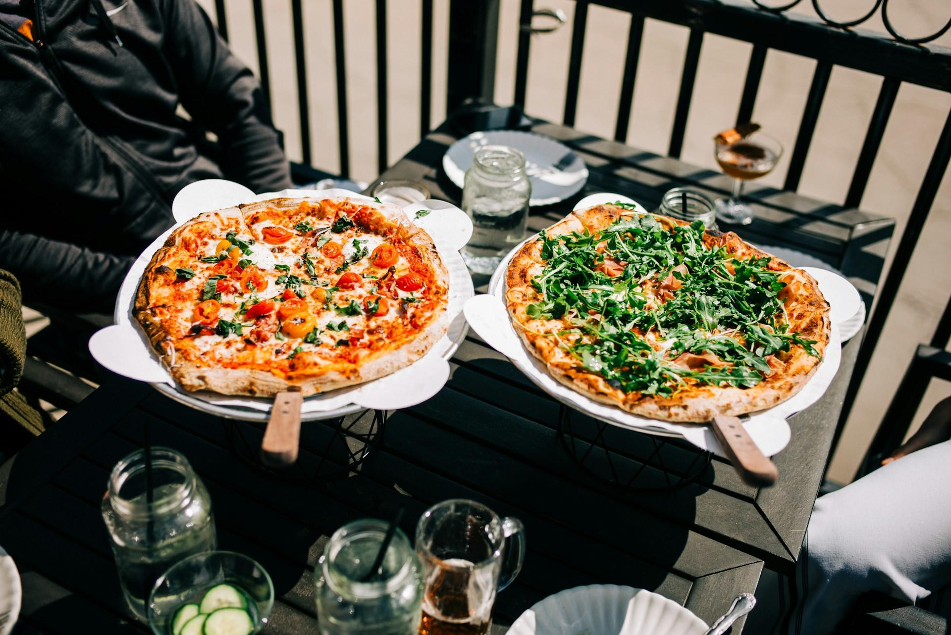 Dos pizzas en una mesa al aire libre con una persona al fondo.