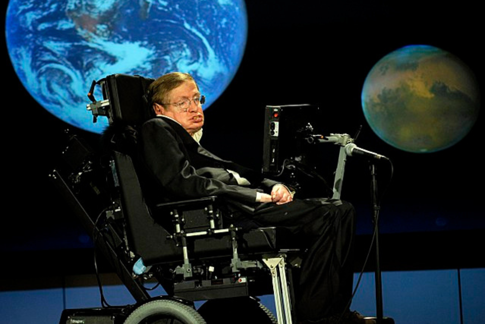 Hawking en silla de ruedas en el escenario con imágenes proyectadas de la Tierra y otro planeta de fondo.