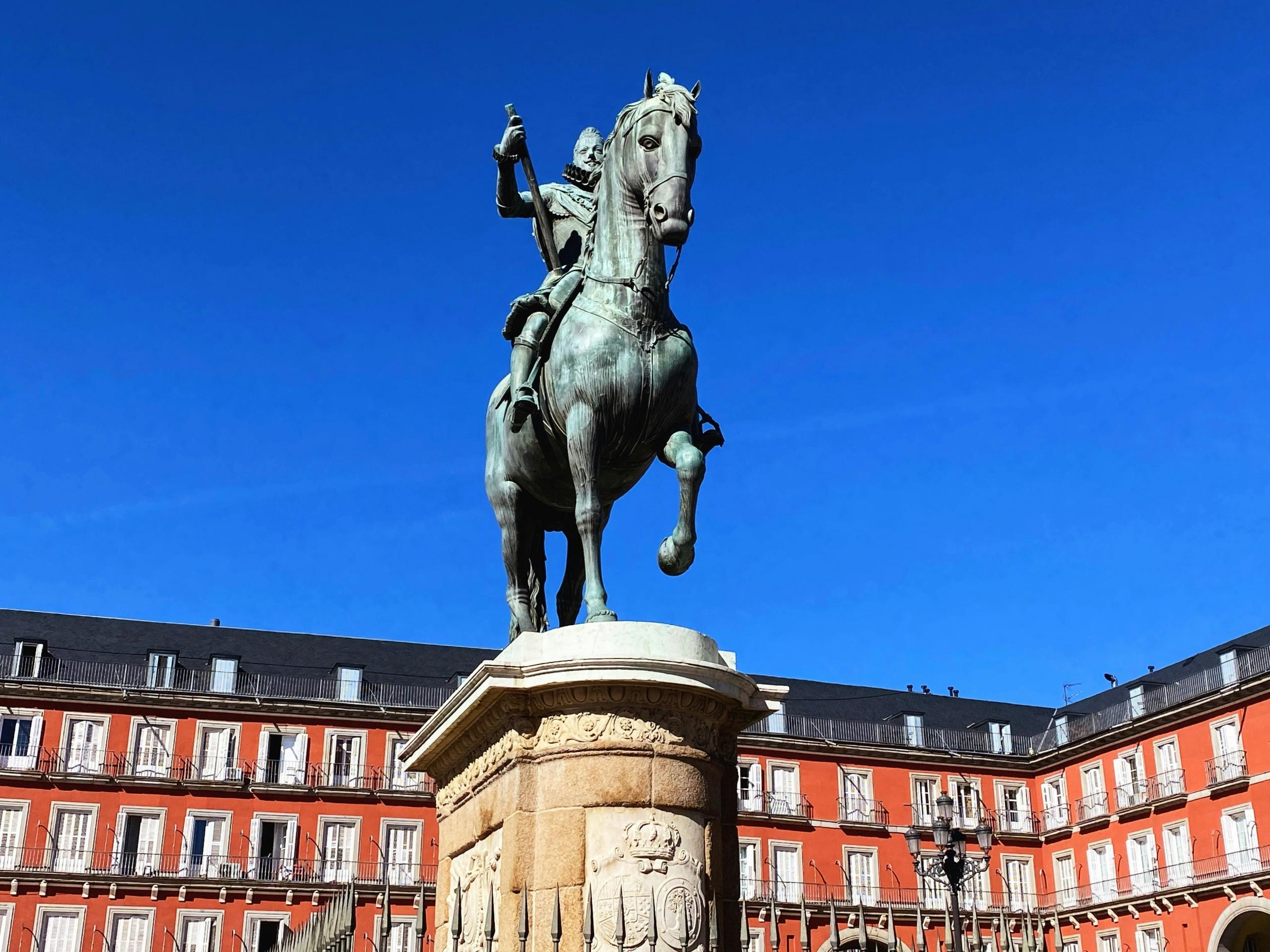 Estatua ecuestre de bronce en una plaza con edificios rojos bajo un cielo azul de la plaza mayor de madrid