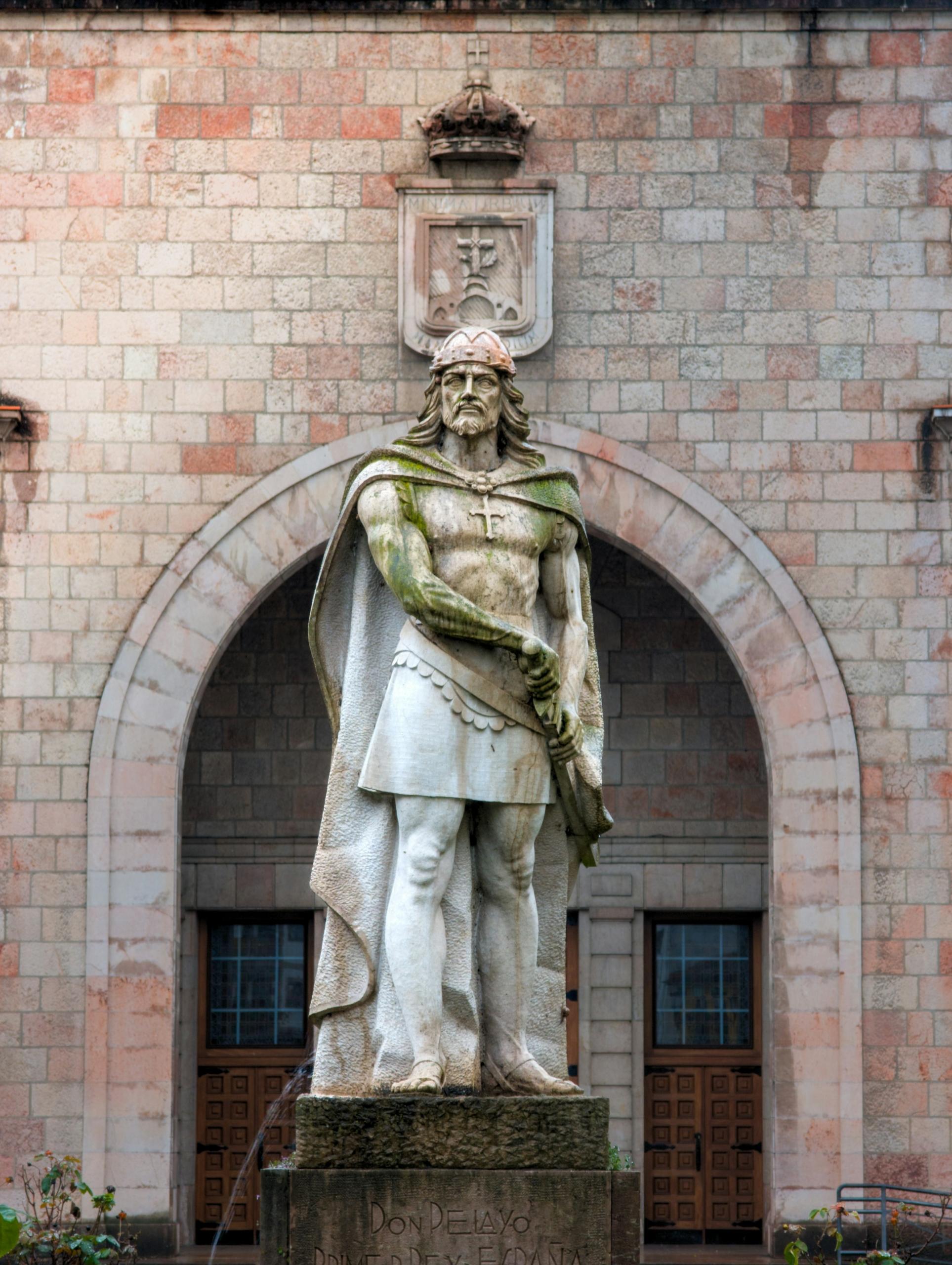 Estatua del personaje histórico pelayo con capa delante de un edificio de piedra arqueado.
