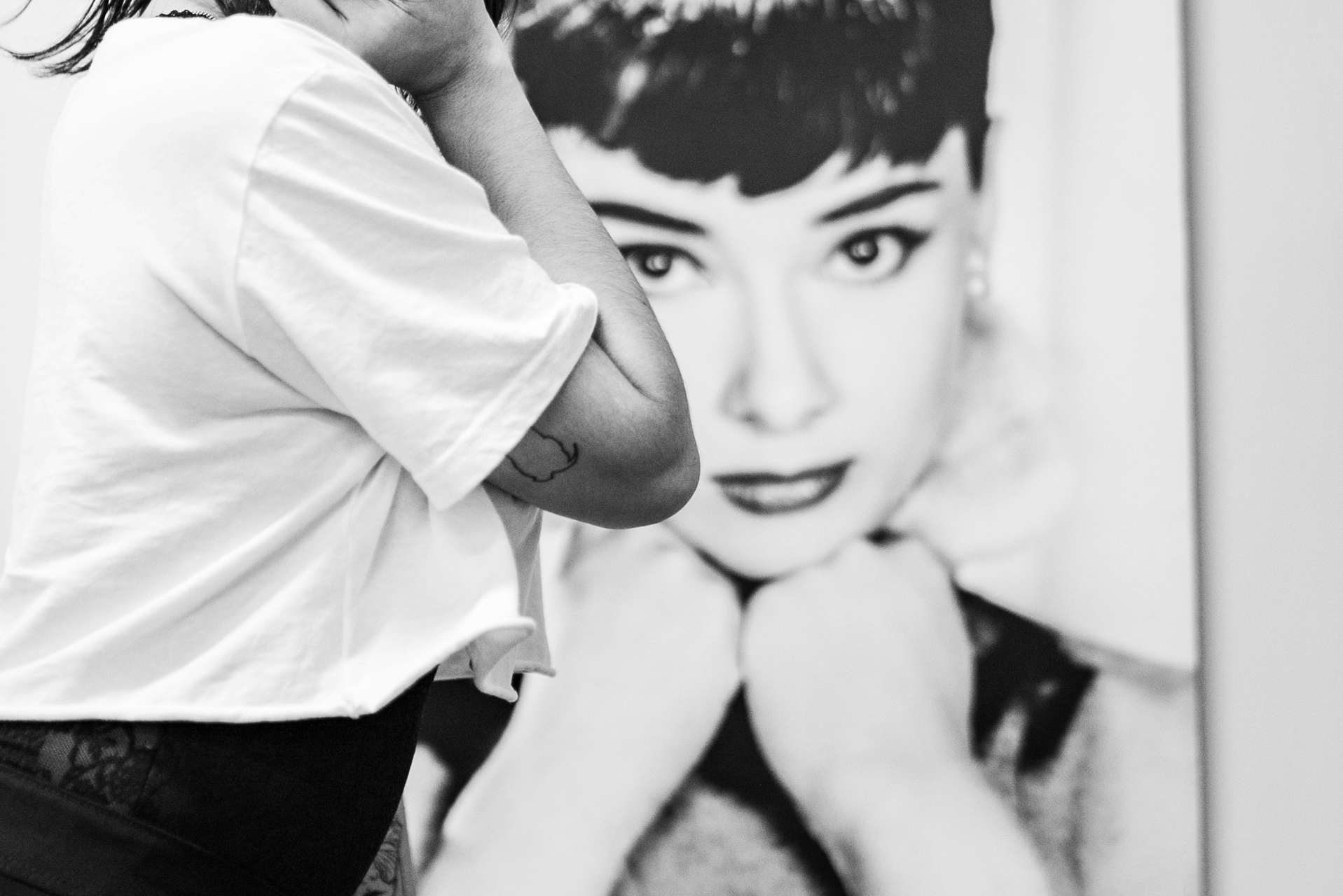 Persona con camiseta blanca de pie con el brazo cruzado, tatuaje visible en el brazo con un poster de Audrey al fondo.
