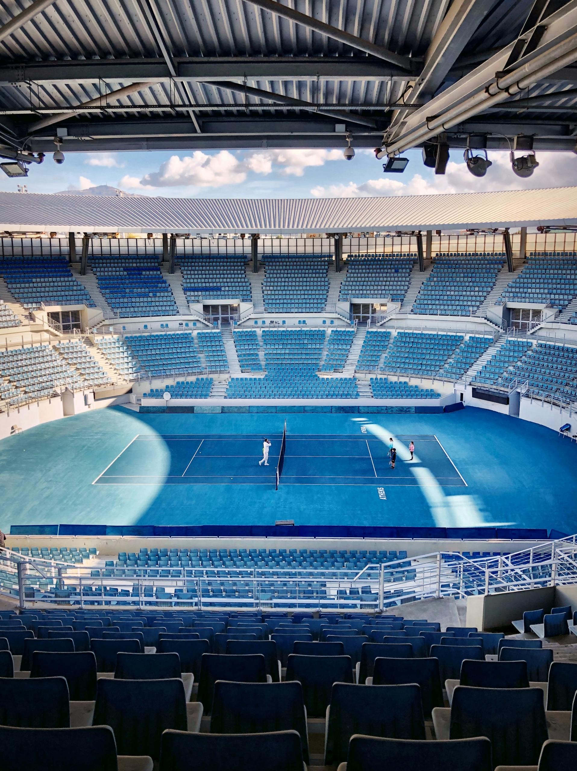 Estadio de tenis vacío con asientos azules y unas pocas personas en la pista bajo un techo abierto.