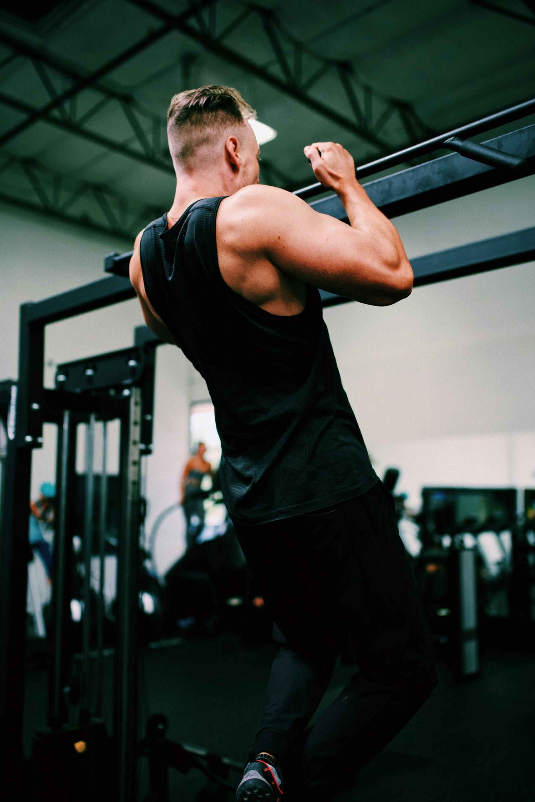 Un hombre con una camiseta sin mangas negra hace una dominada en un gimnasio. Sus brazos musculosos destacan, transmitiendo fuerza y concentración. Al fondo se ven máquinas de gimnasio.