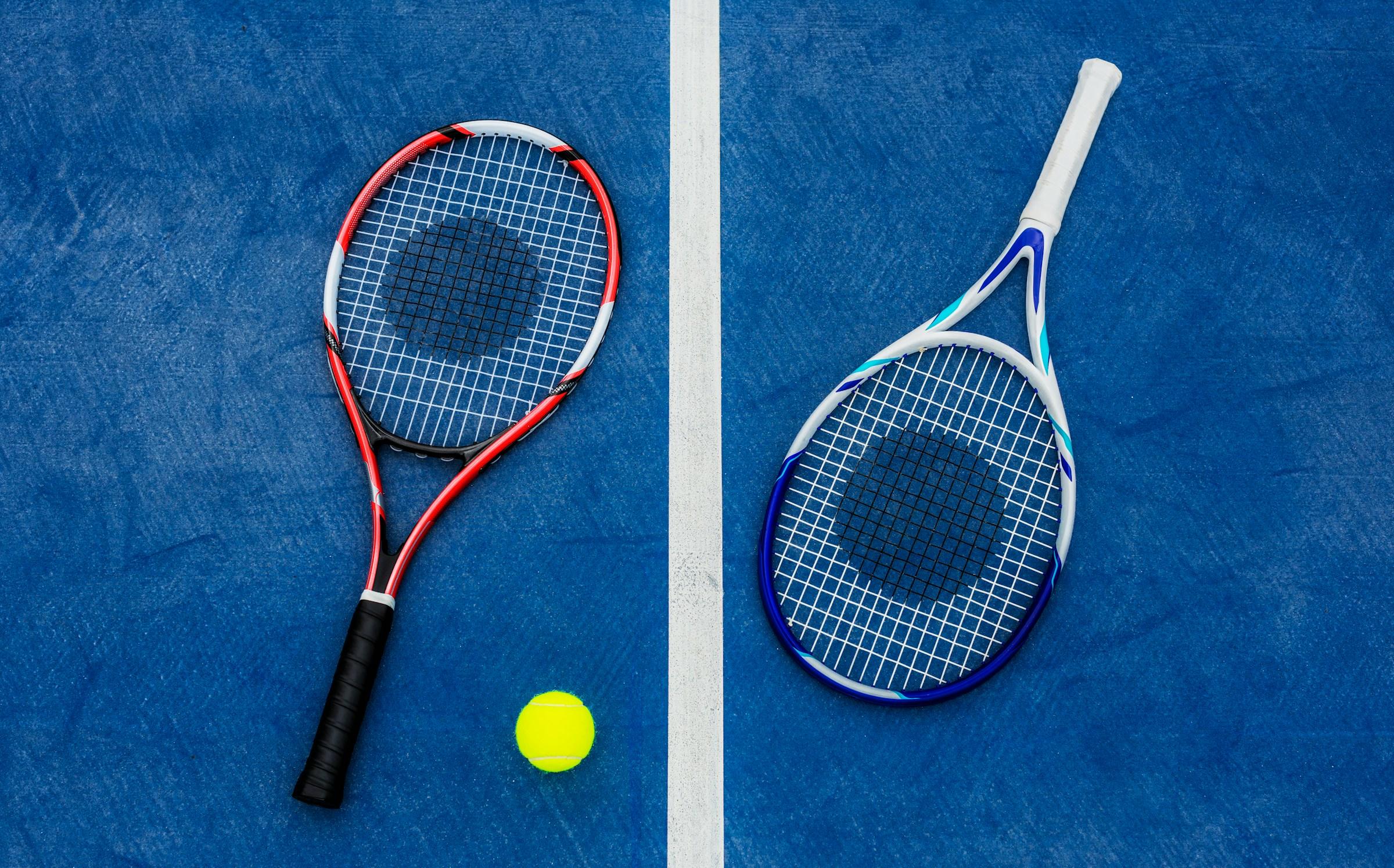 Dos raquetas de tenis y una pelota en una pista azul con línea blanca.