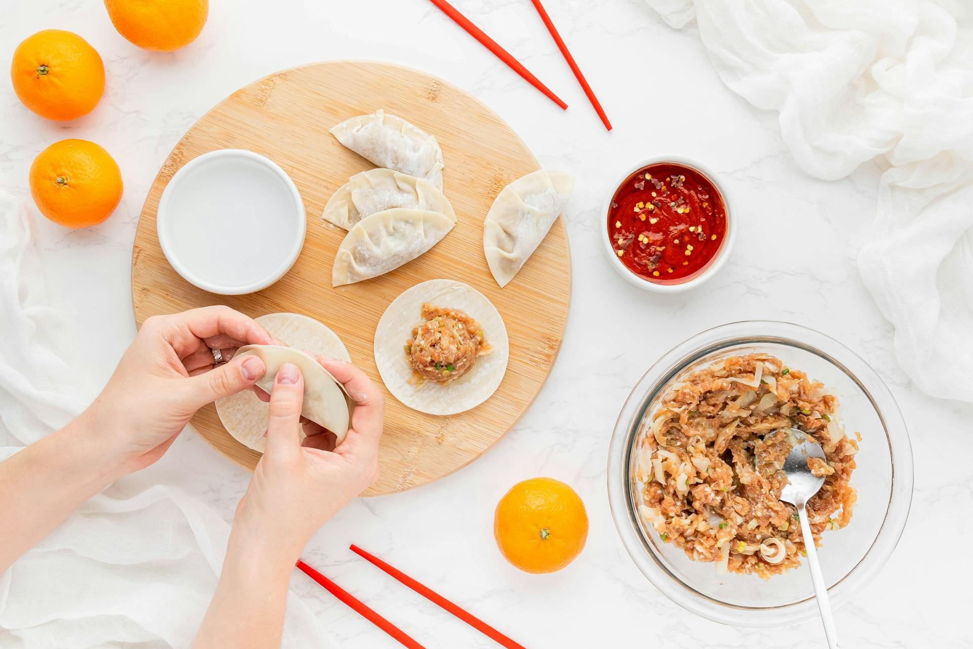 Manos haciendo dumplings junto a ingredientes y salsa sobre una superficie blanca.