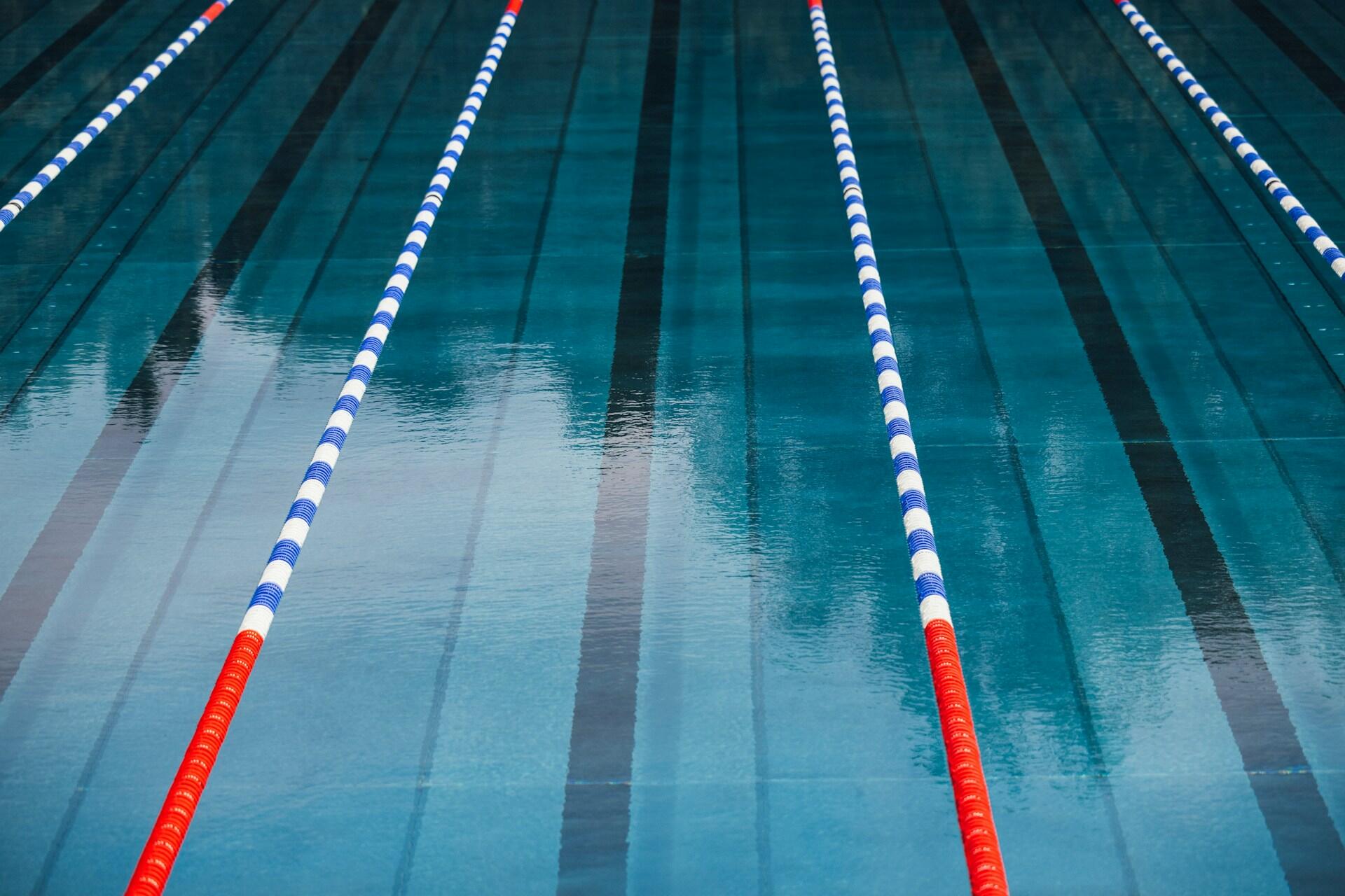 Una piscina tranquila con cinco carriles delimitados por separadores flotantes de color azul, blanco y rojo. El agua, que refleja la luz, crea un ambiente sereno y ordenado.