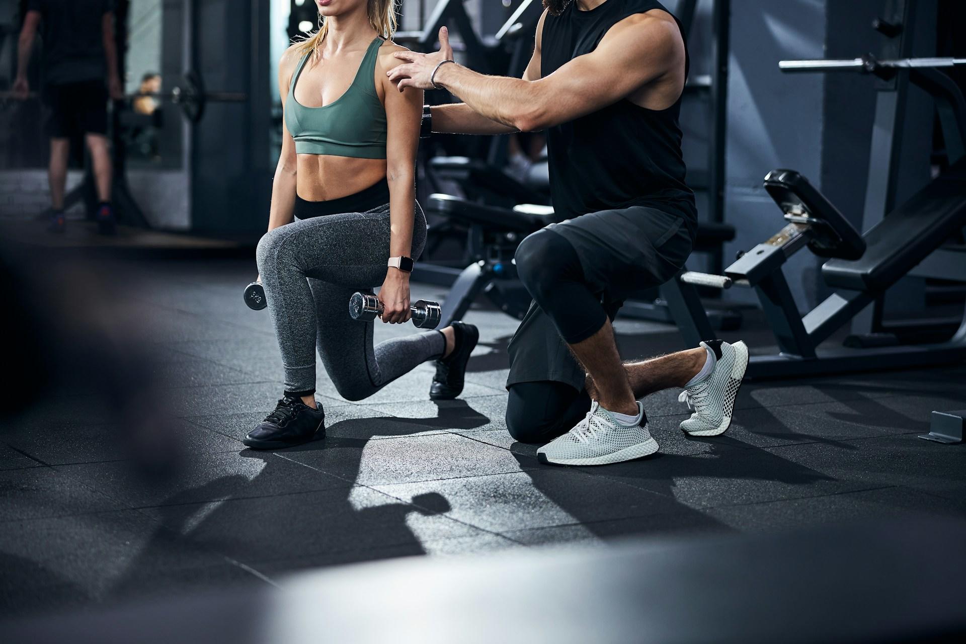 Una mujer realiza una estocada con mancuernas, guiada por un entrenador masculino en un gimnasio.