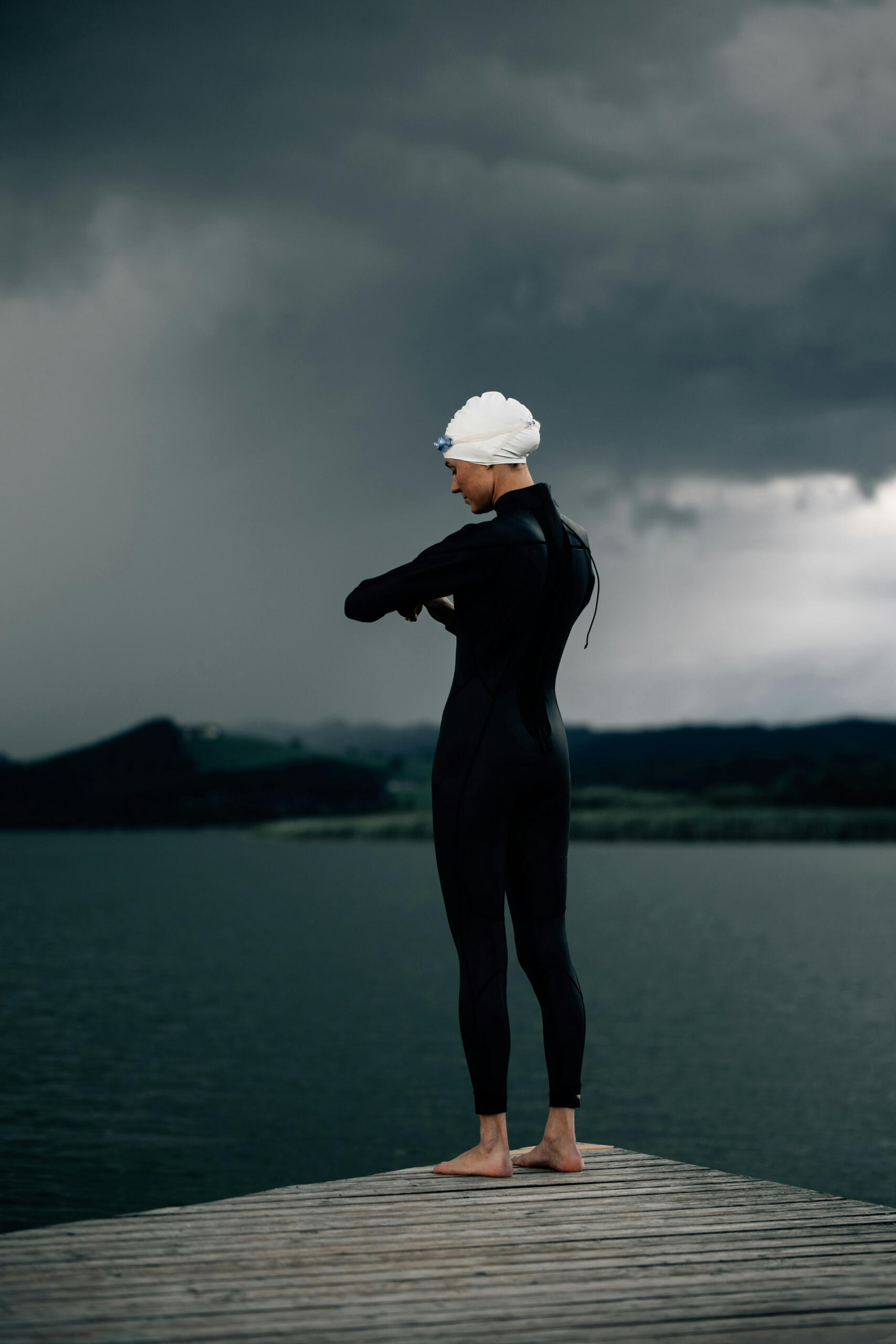 Persona con traje de neopreno y gorro de baño en un muelle con vistas a un lago tormentoso.