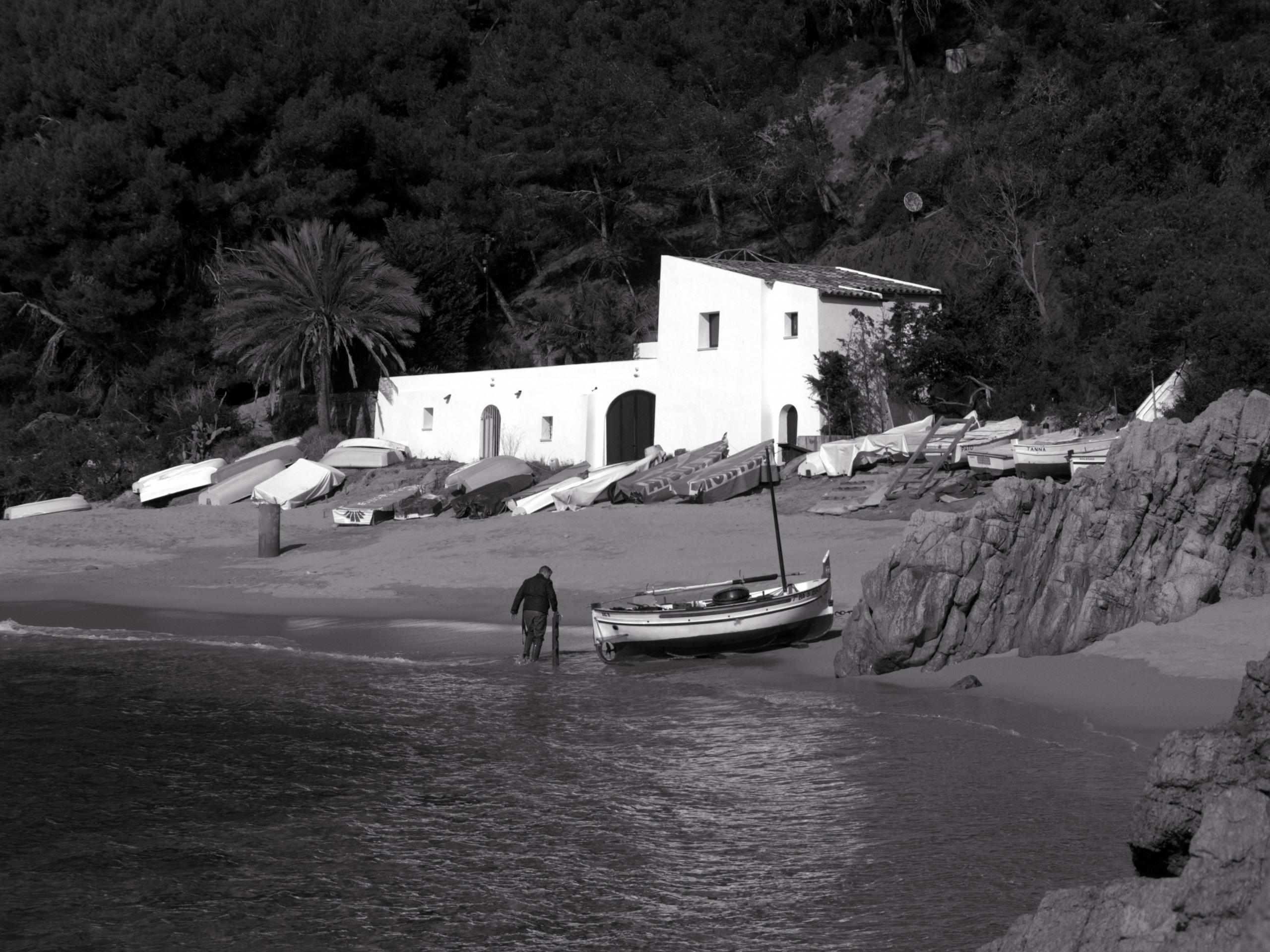 Imagen monocroma de una persona junto al mar, cerca de barcos y un edificio blanco, con árboles al fondo en la costa de girona.