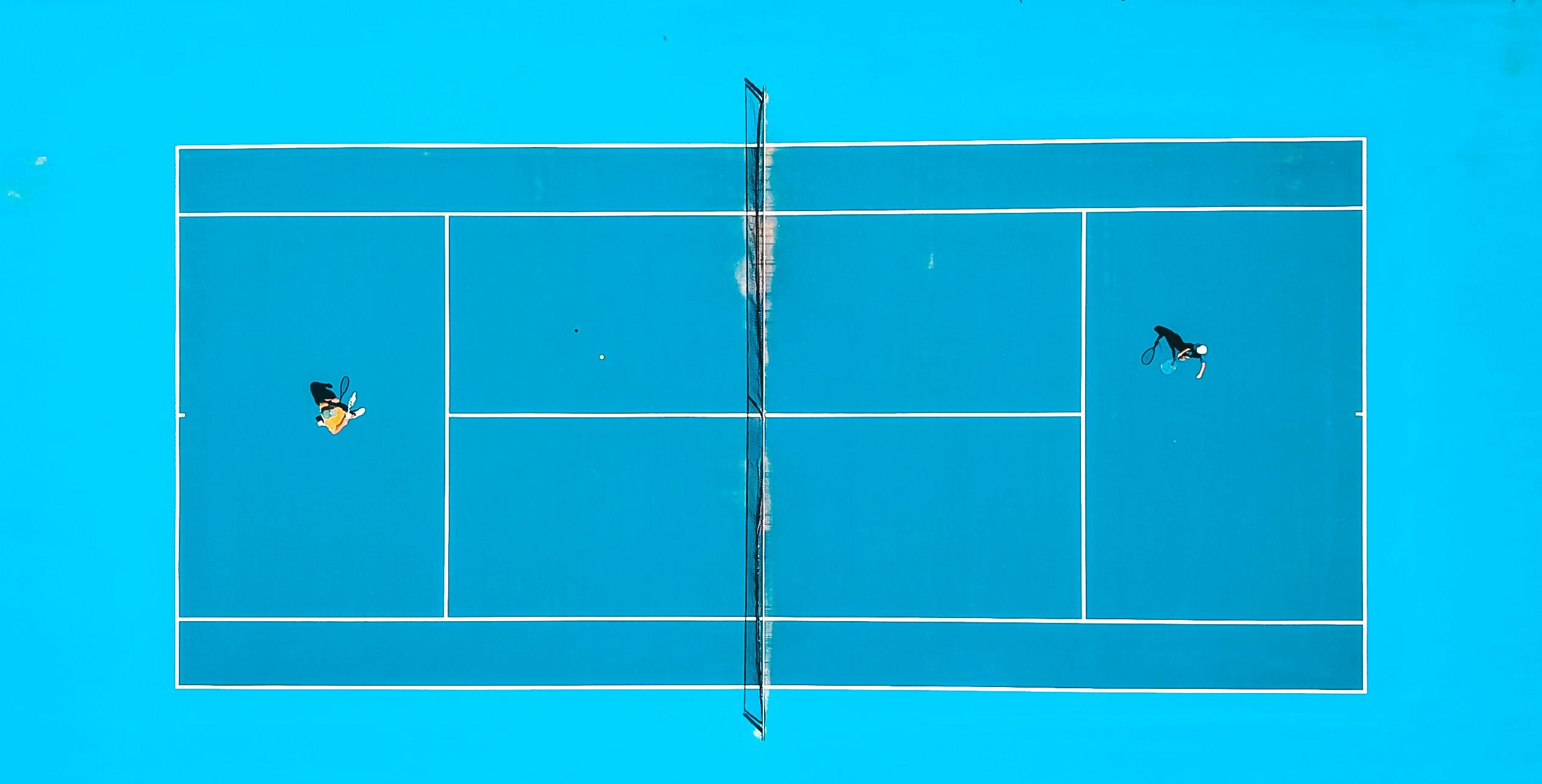 Vista aérea de dos jugadores en una pista de tenis azul dividida por una red.