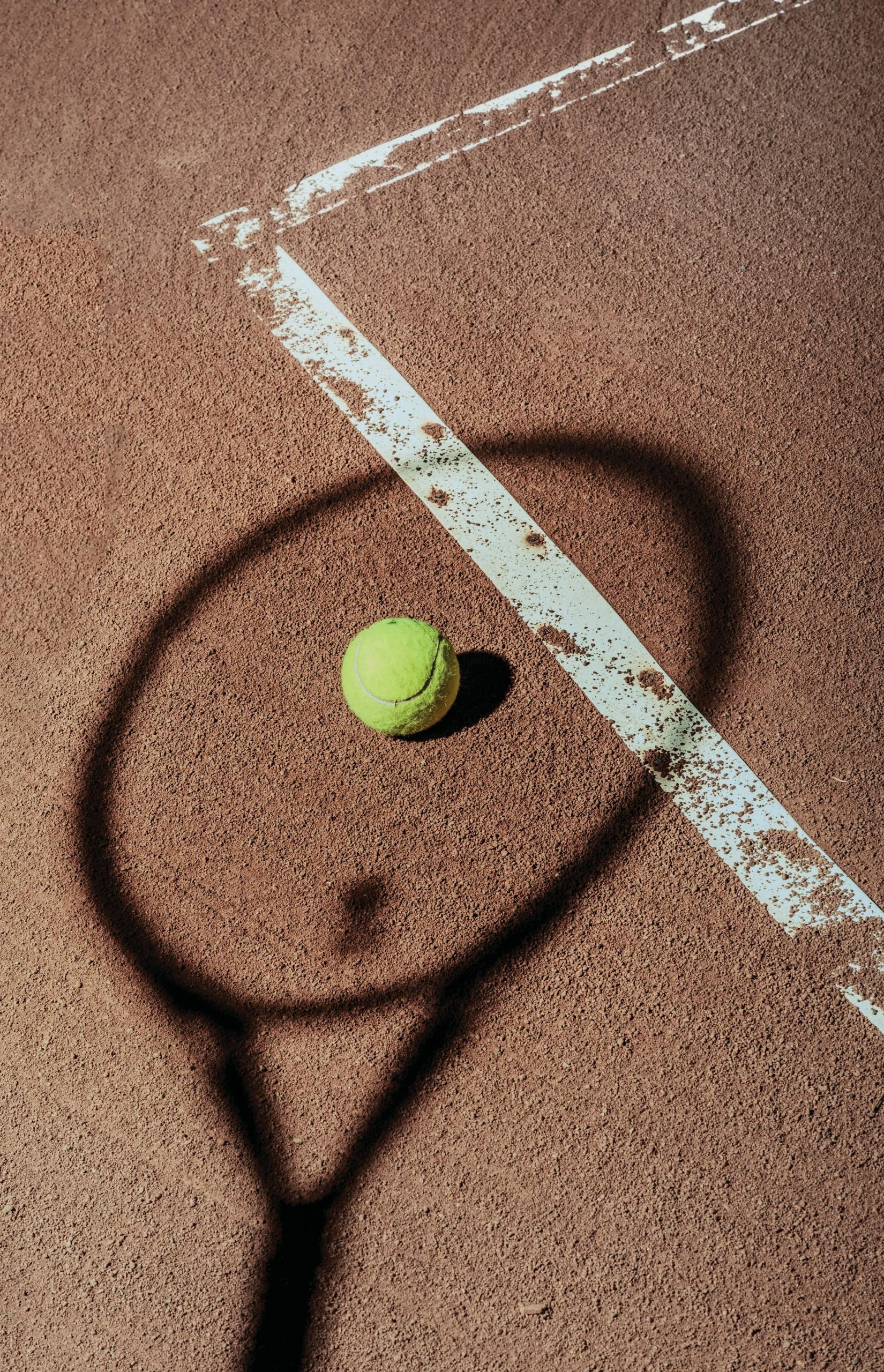 Pelota de tenis en pista de tierra batida cerca de línea blanca con sombra visible de raqueta de tenis.