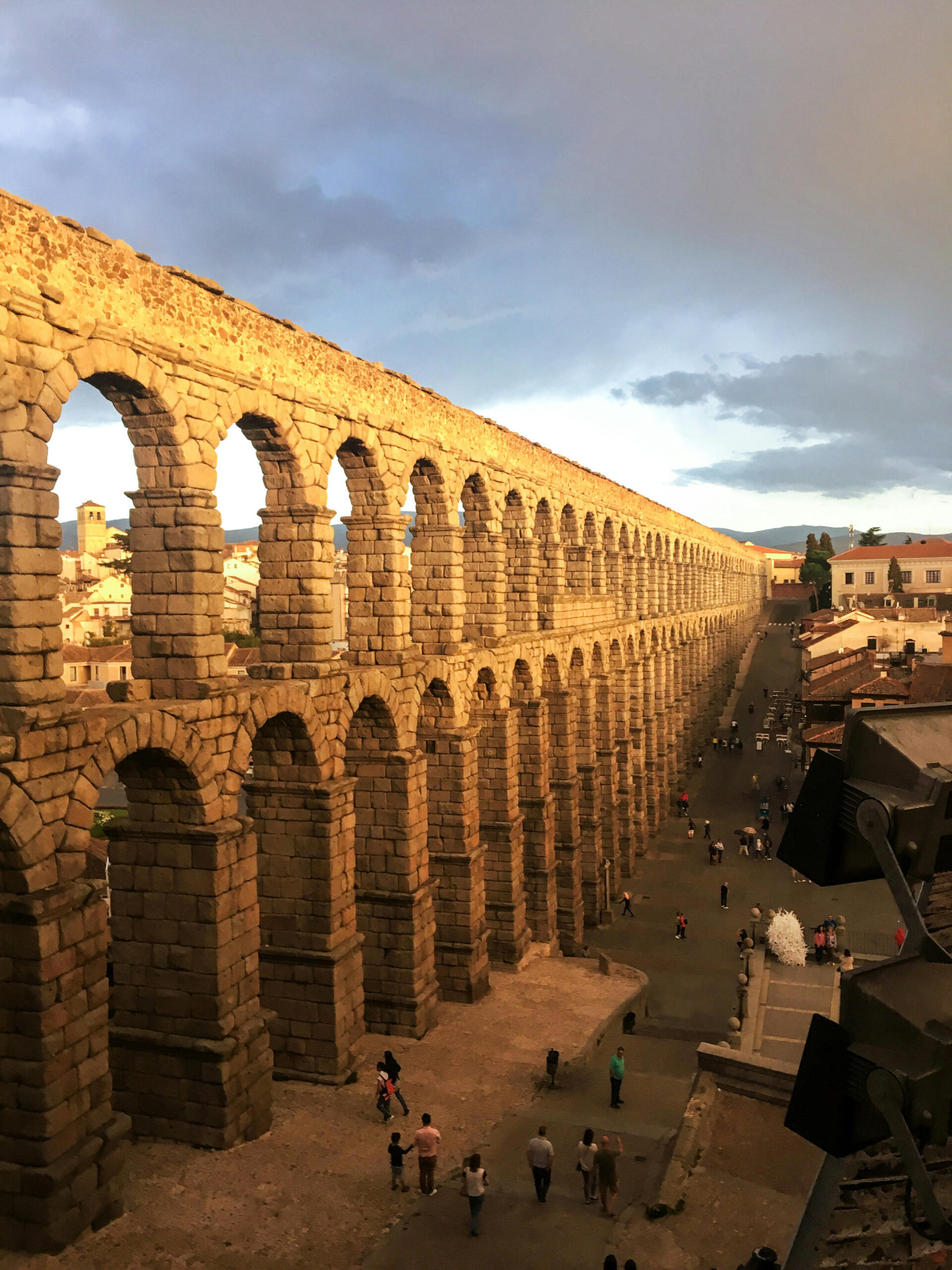 Antiguo acueducto con arcos dobles al atardecer, proyectando sombras sobre una calle de la ciudad con gente paseando.