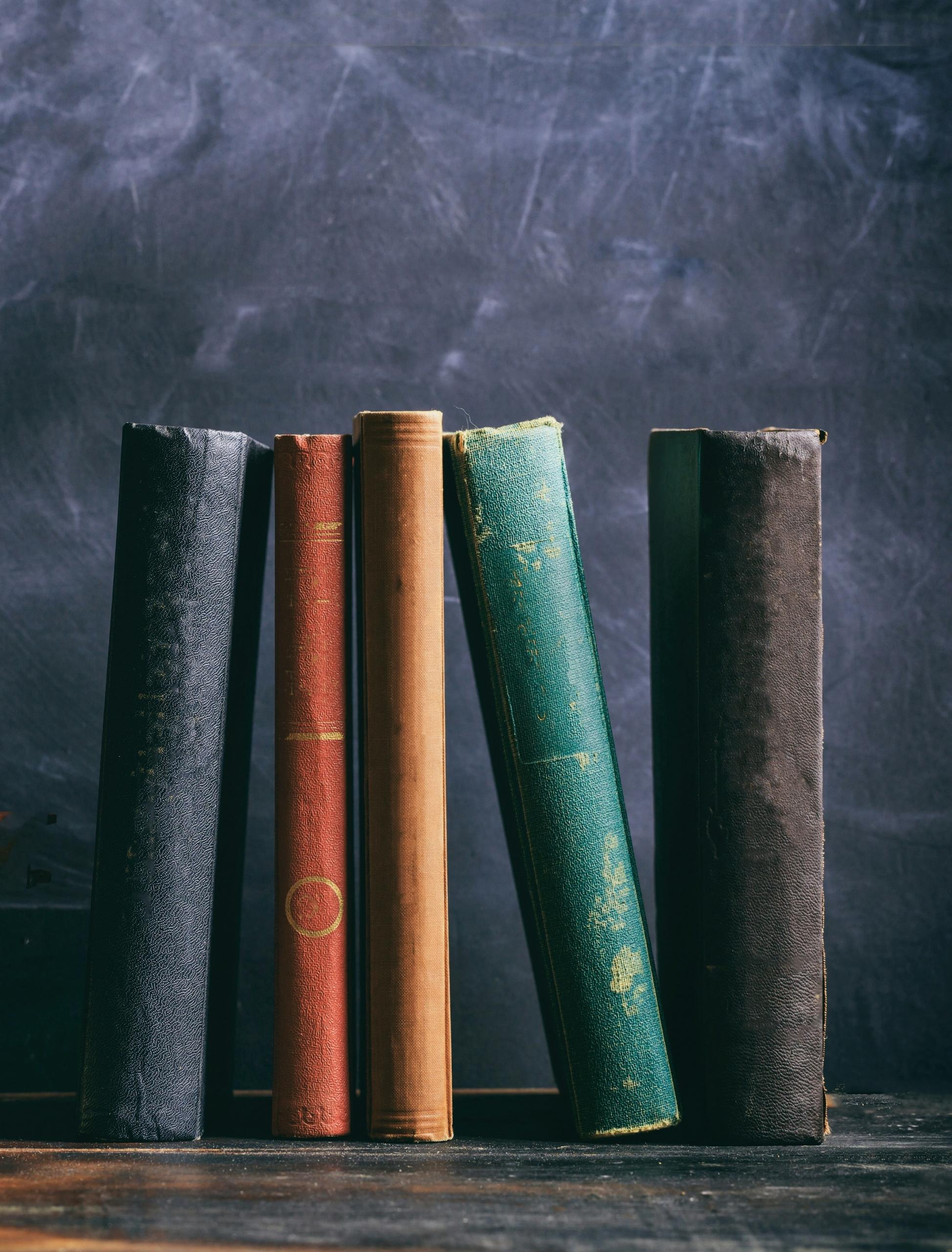 Cinco libros antiguos en posición vertical sobre una superficie de madera con un fondo oscuro y texturado.