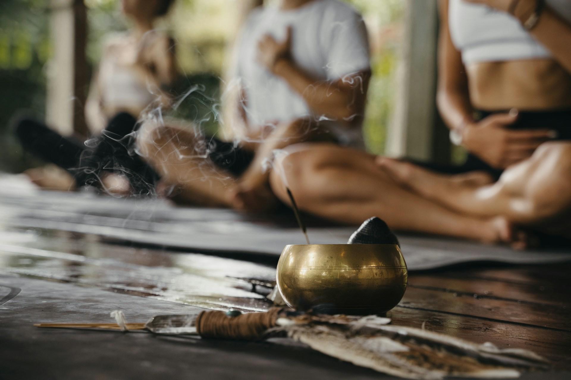 Un cuenco tibetano con humo de incienso, delante de personas borrosas en postura de meditación.