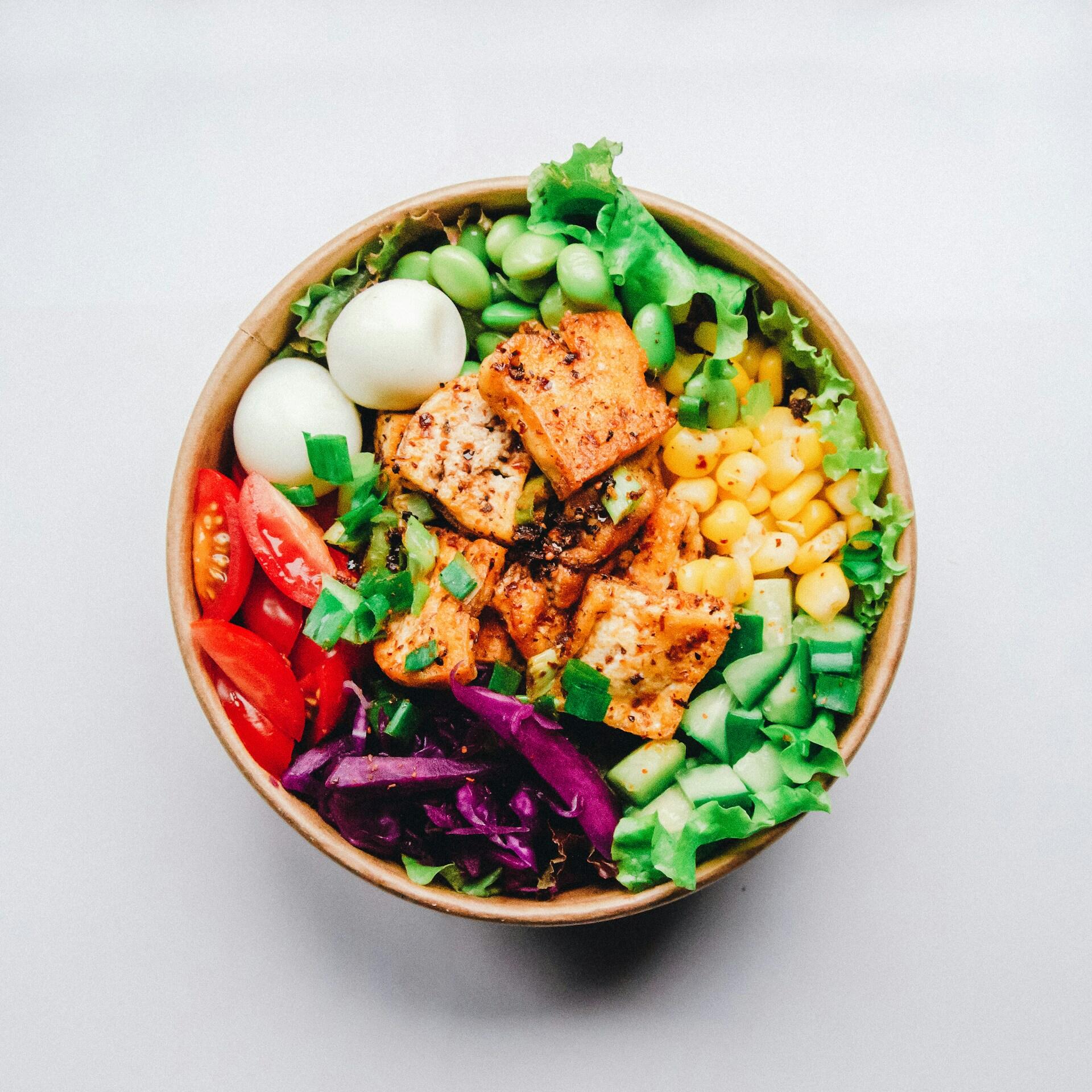 Un colorido cuenco de ensalada con tofu a la plancha, tomates cherry, maíz, pepinos, edamame, huevos cocidos y col morada, sobre un fondo blanco.