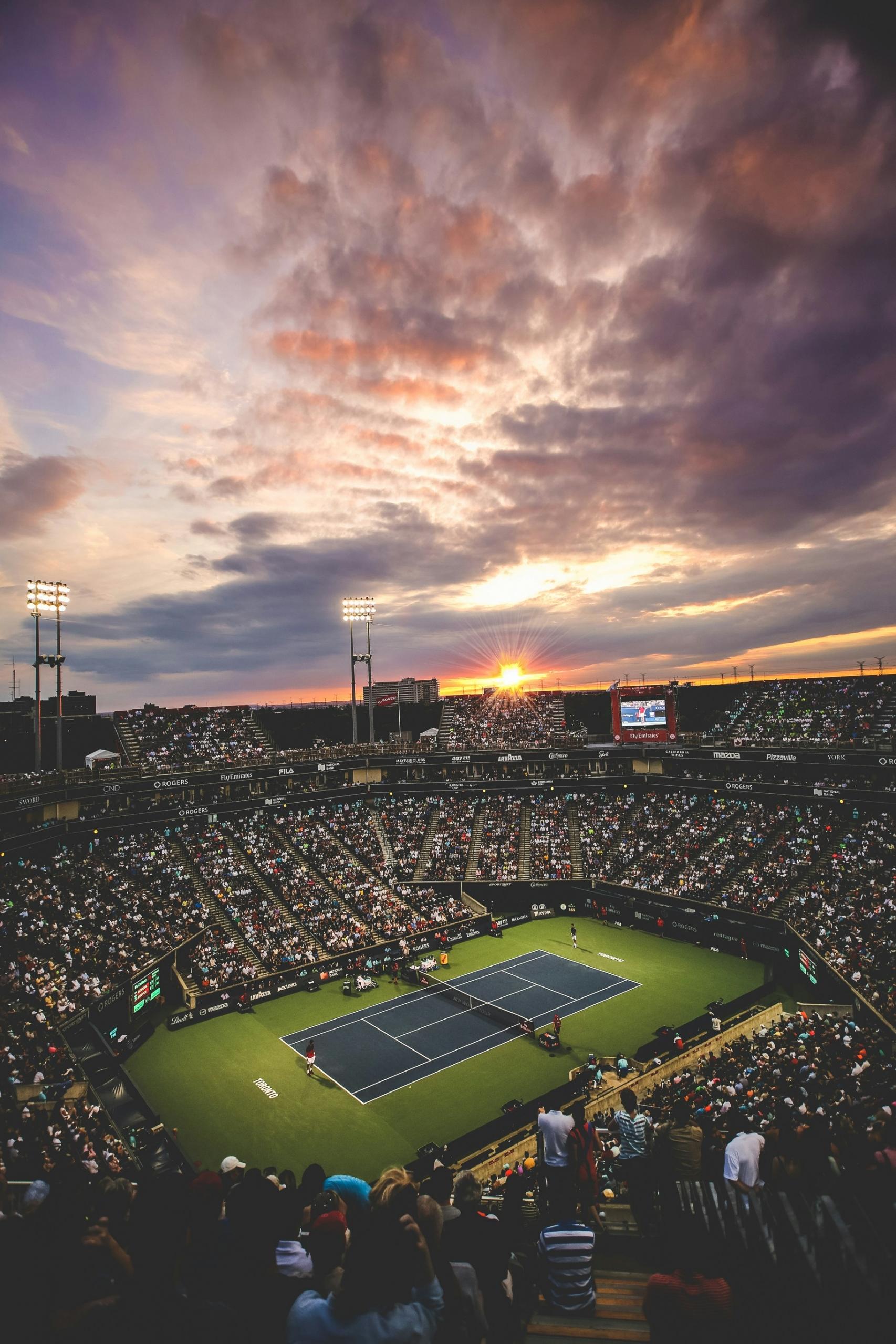Puesta de sol sobre un estadio de tenis con un público repleto y nubes espectaculares en el cielo.