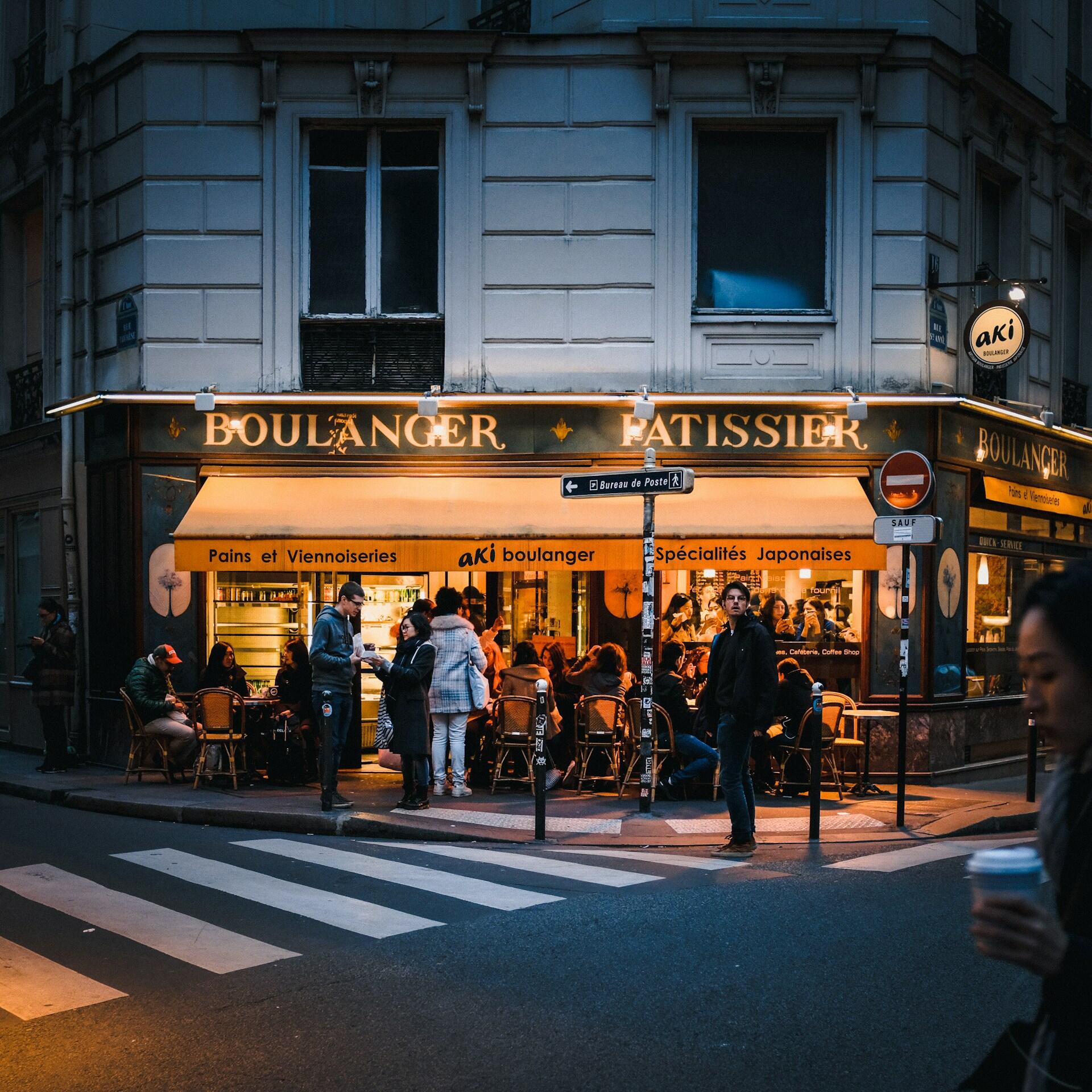Una acogedora panadería parisina al atardecer, con una cálida iluminación, y clientes sentados en las mesas de la terraza. En primer plano, una persona cruza la calle con un café en la mano.