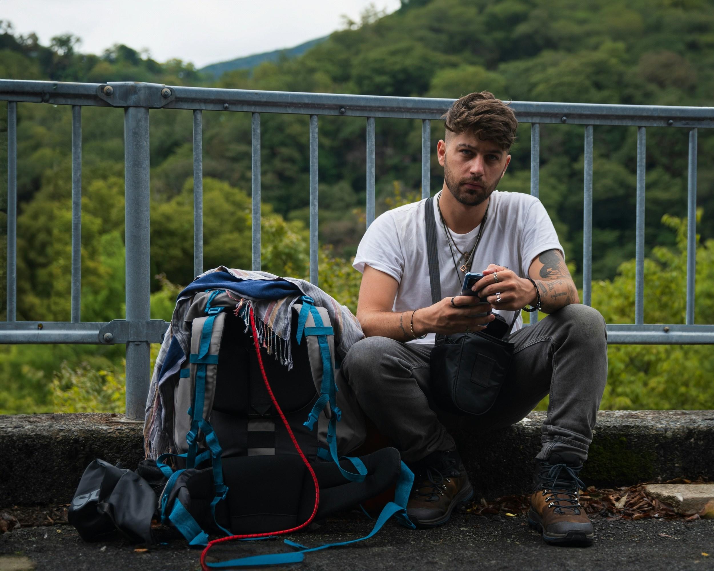 Una persona sentada junto a una barandilla con una mochila, utilizando un smartphone.