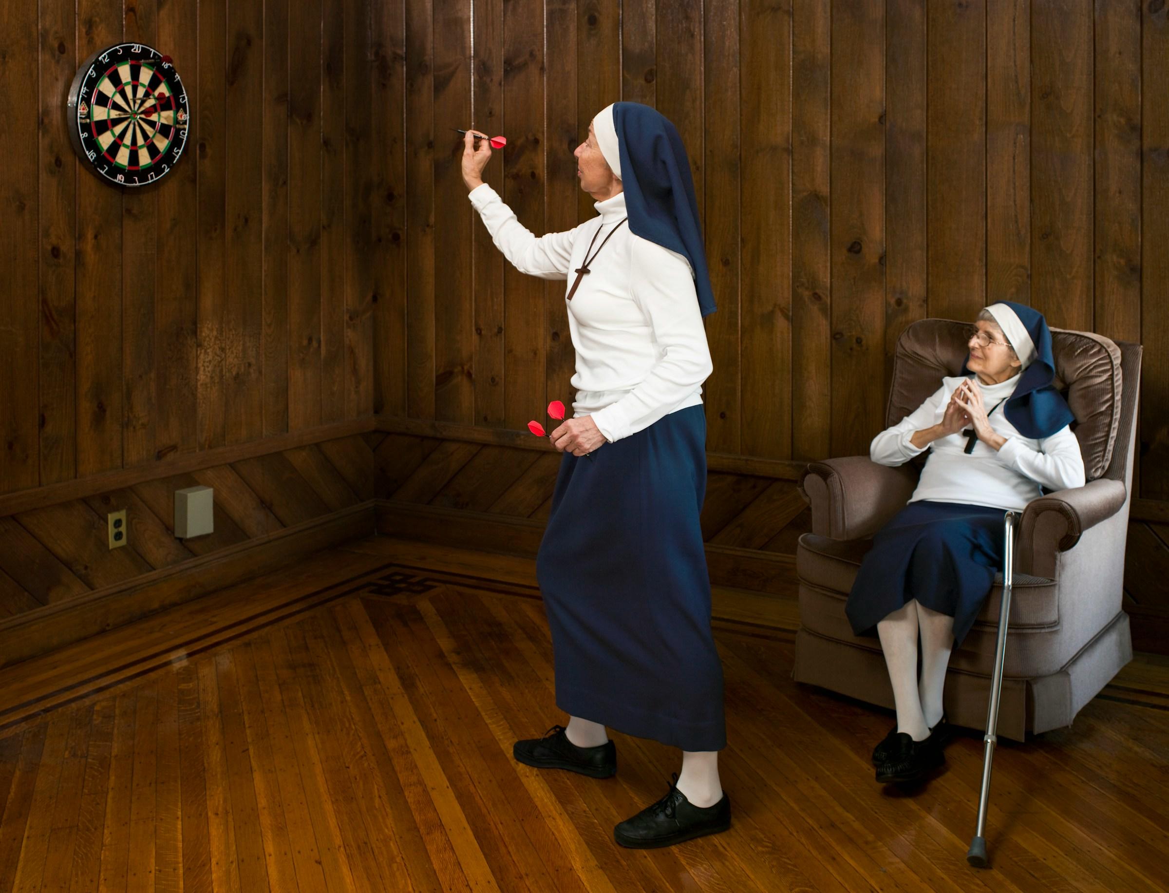 Dos monjas en una habitación con paneles de madera, una de pie y jugando a los dardos, la otra sentada con las manos juntas.