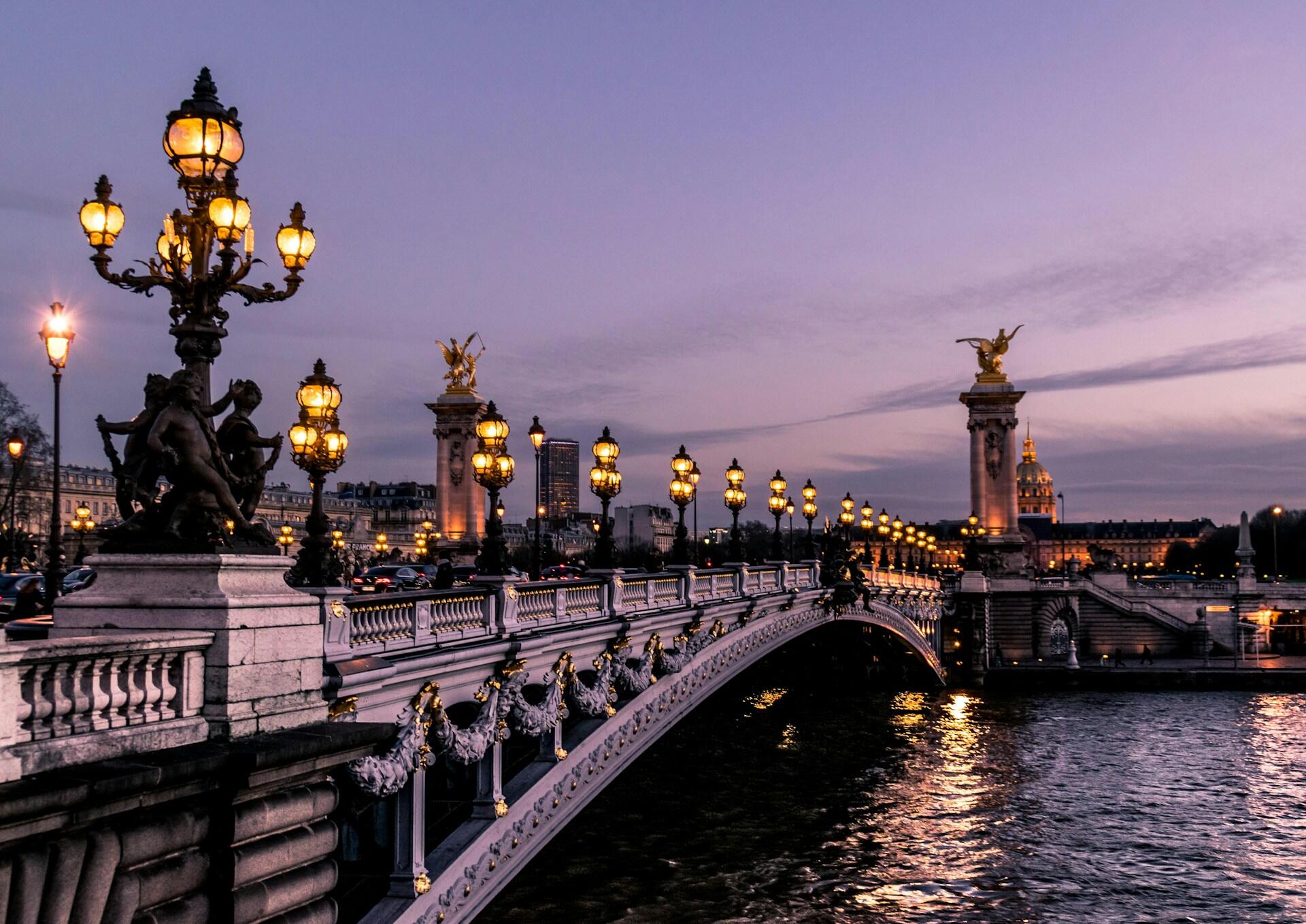 Al caer la tarde, las farolas iluminan el ornamentado puente Alexandre III de París, con sus estatuas doradas en lo alto de los pilares. Su reflejo brilla en el río que discurre a sus pies.