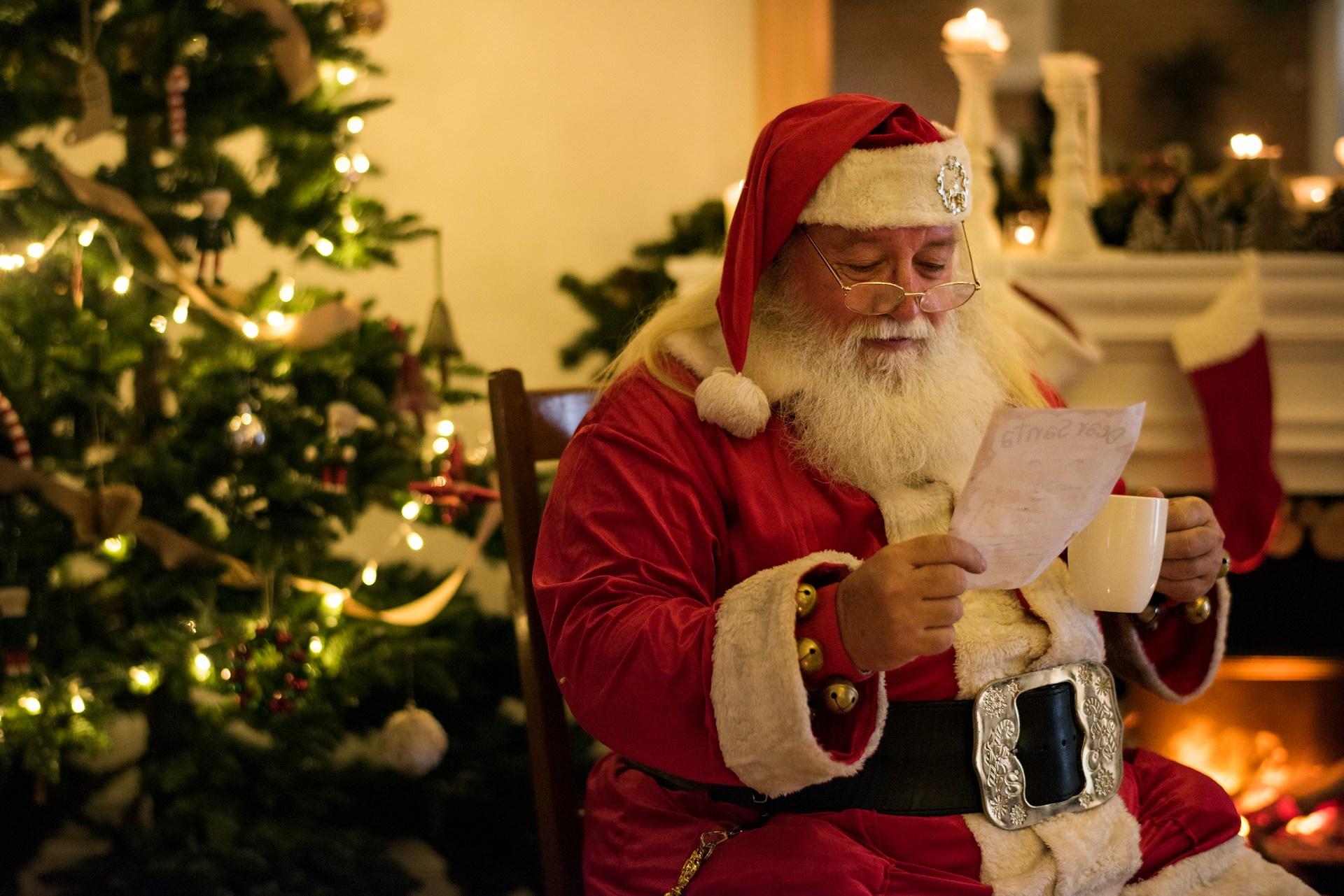 Persona disfrazada de Papá Noel sosteniendo una lista y una taza, con un árbol de Navidad decorado al fondo.