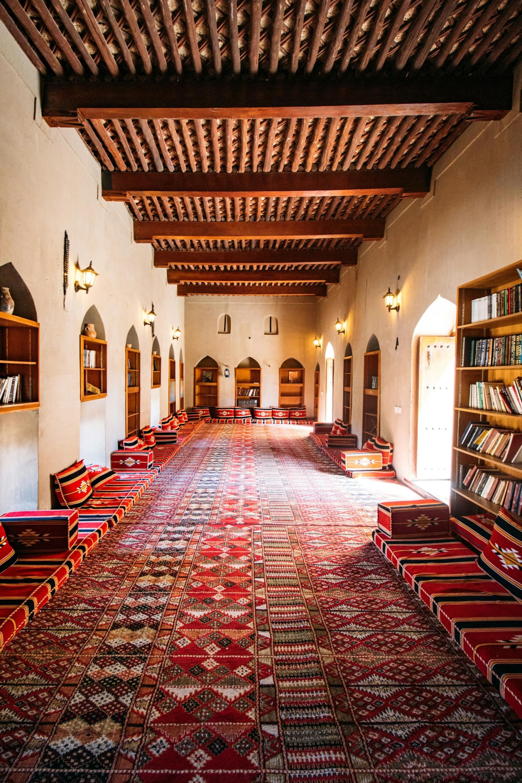 Sala de lectura histórica con vigas de madera ornamentadas, alfombras con motivos coloridos, bancos acolchados, estanterías y lámparas que desprenden una luz cálida, creando un ambiente acogedor.