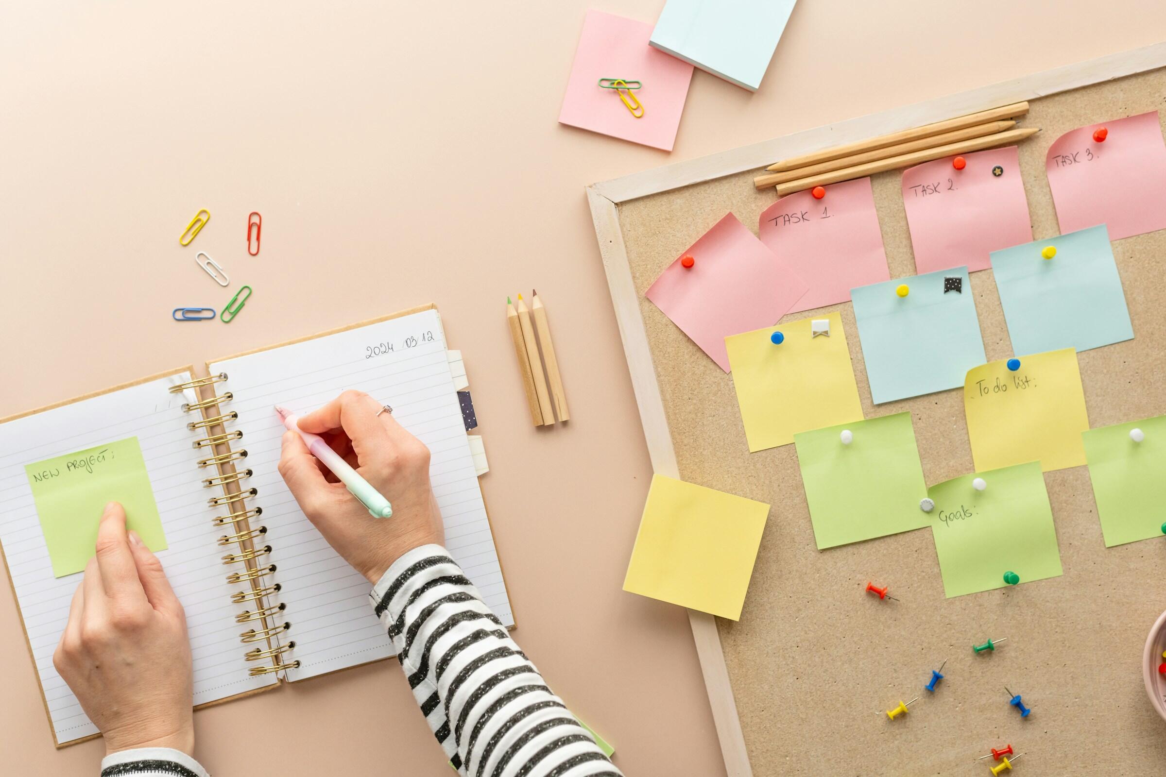 Manos escribiendo en una agenda junto a notas adhesivas de colores en una pizarra de corcho, con lápices y clips.
