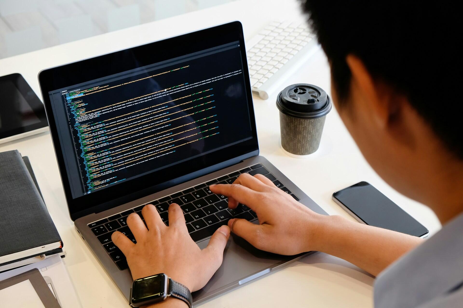 Persona escribiendo en un portátil con un código en la pantalla, un smartphone y una taza de café sobre la mesa.