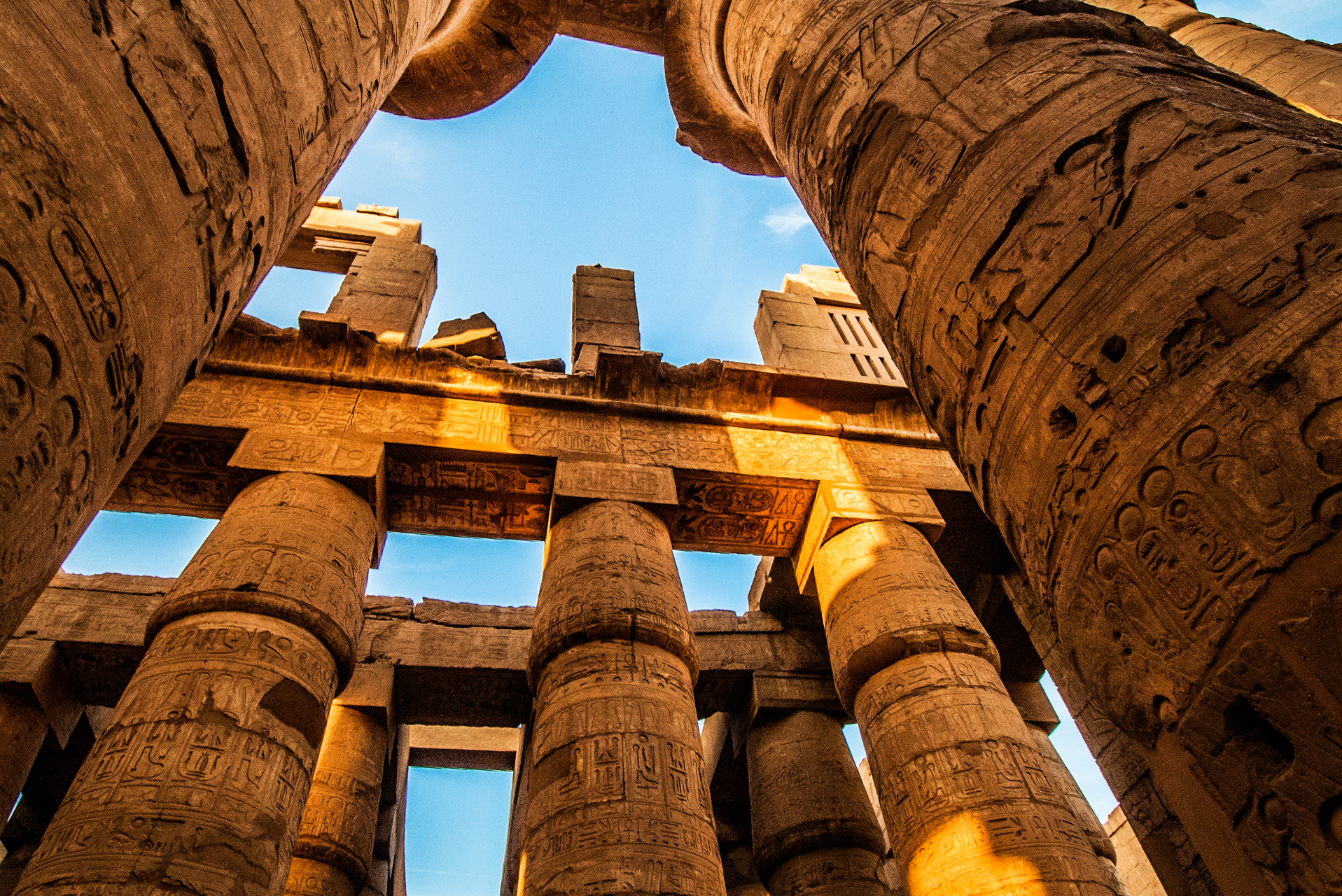 Vista desde abajo de enormes columnas de piedra grabadas con jeroglíficos en un antiguo templo egipcio. La luz del sol ilumina parte de las columnas y las estructuras de piedra, mientras el cielo azul se ve de fondo a través de las aberturas en la parte superior.