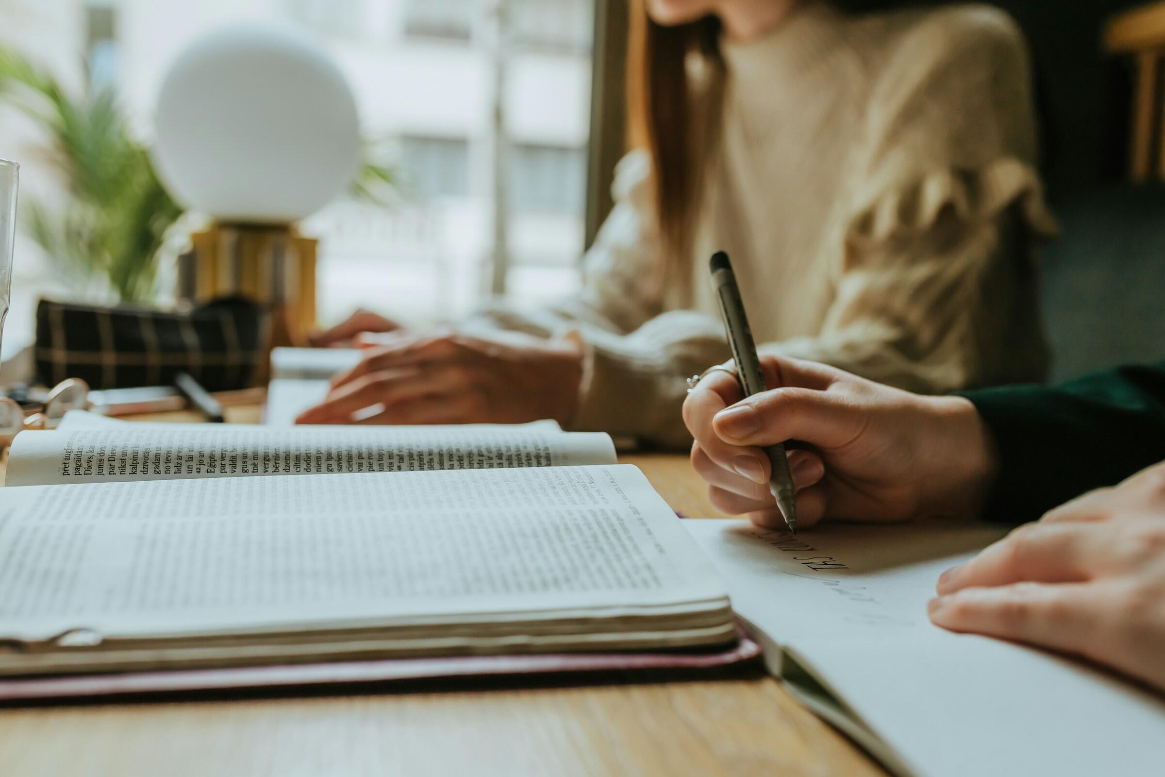 Dos personas estudiando con libros y tomando apuntes sobre una mesa de madera.