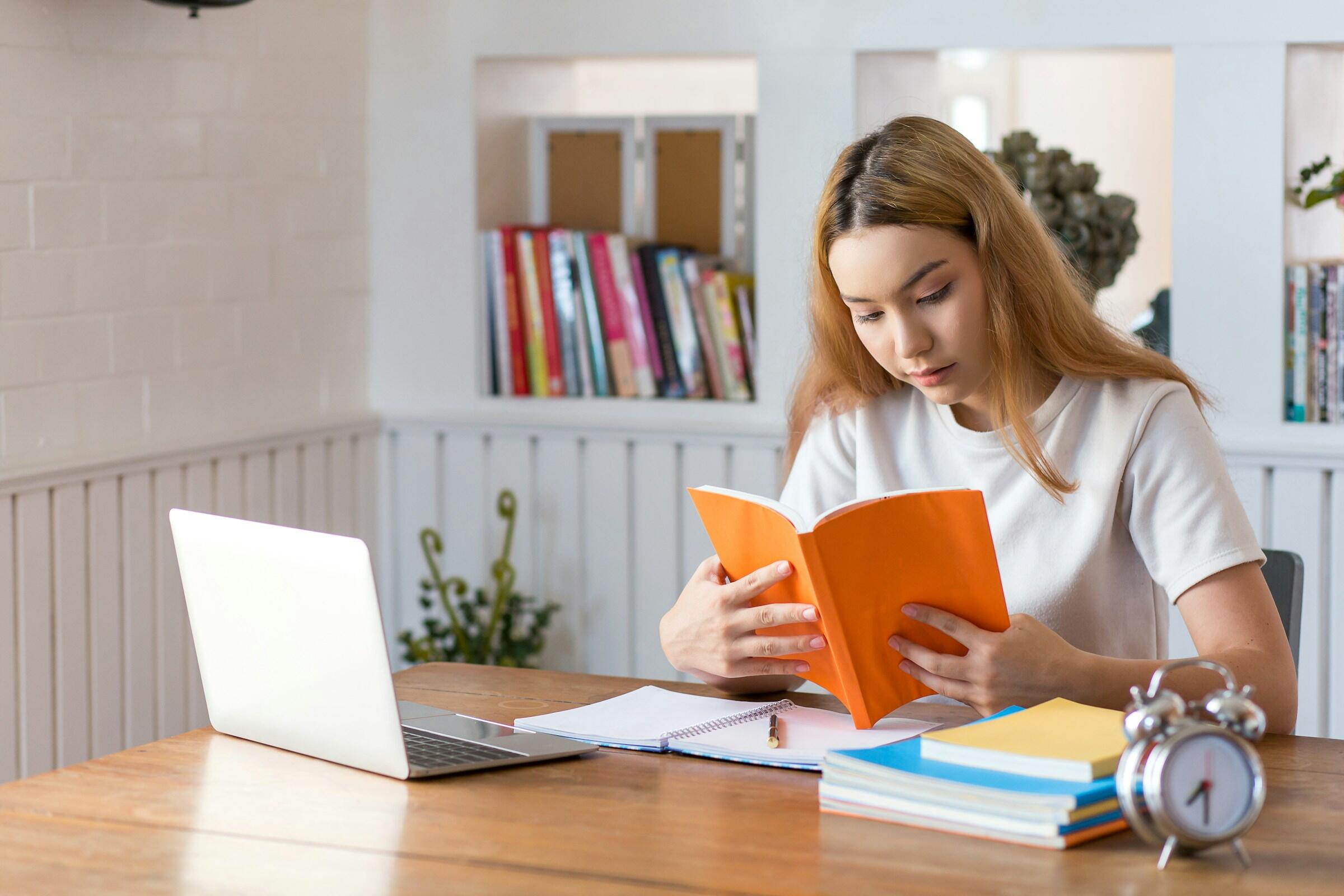 Persona leyendo un libro en un escritorio con ordenador portátil, cuadernos y un reloj.