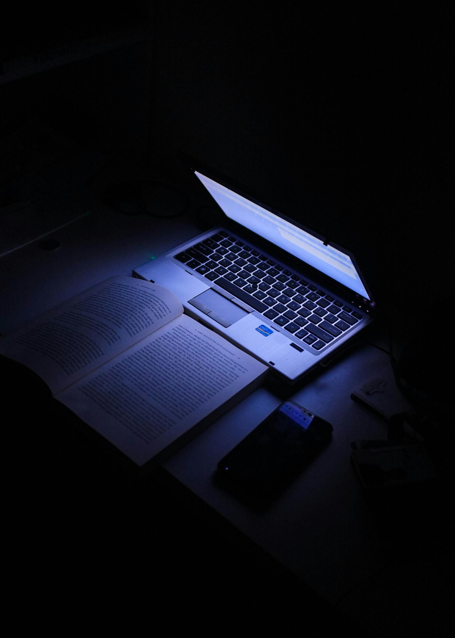 Ordenador portátil iluminando un libro y un smartphone en una habitación oscura.