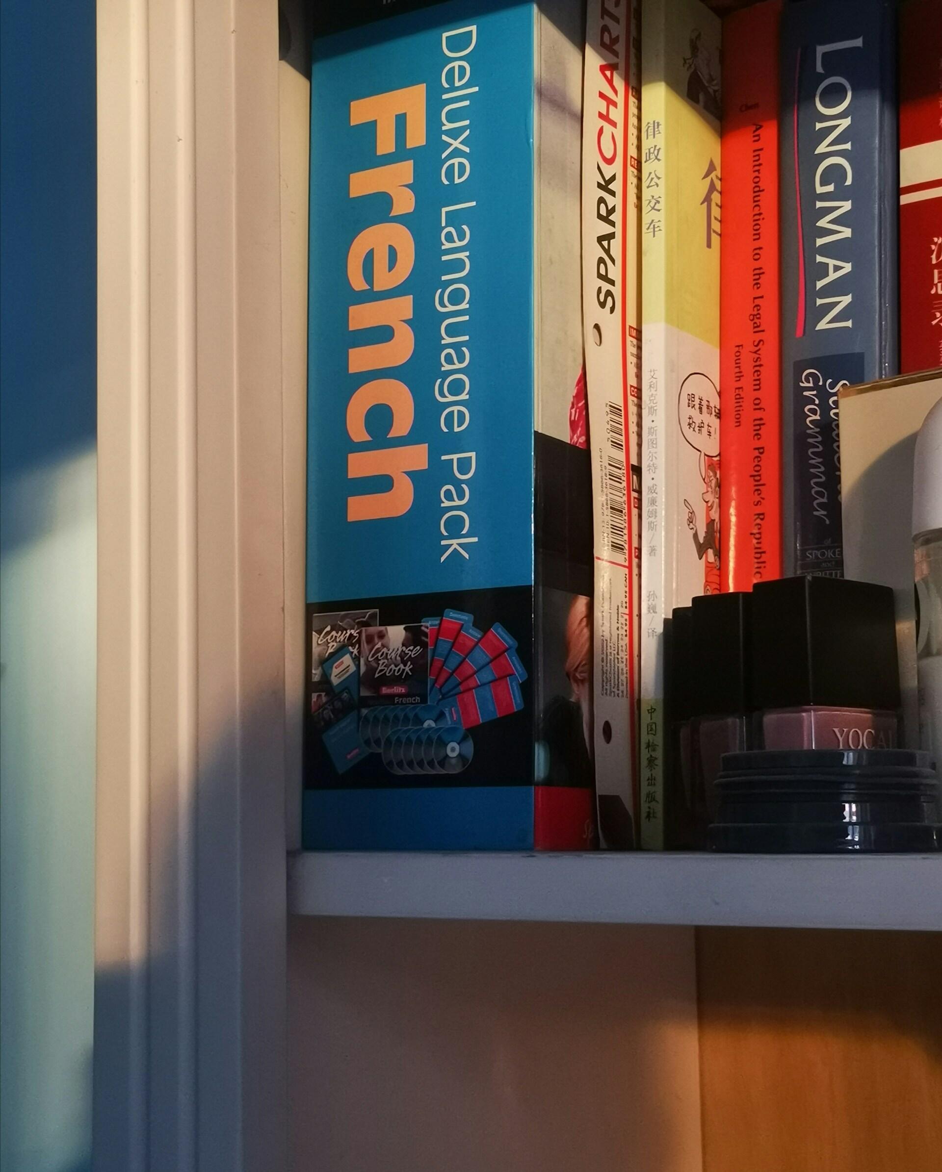 Estantería con diversos libros, entre los que destaca un «Deluxe Language Pack French» de color azul. La suave luz del sol proyecta sombras cálidas, creando un ambiente acogedor.
