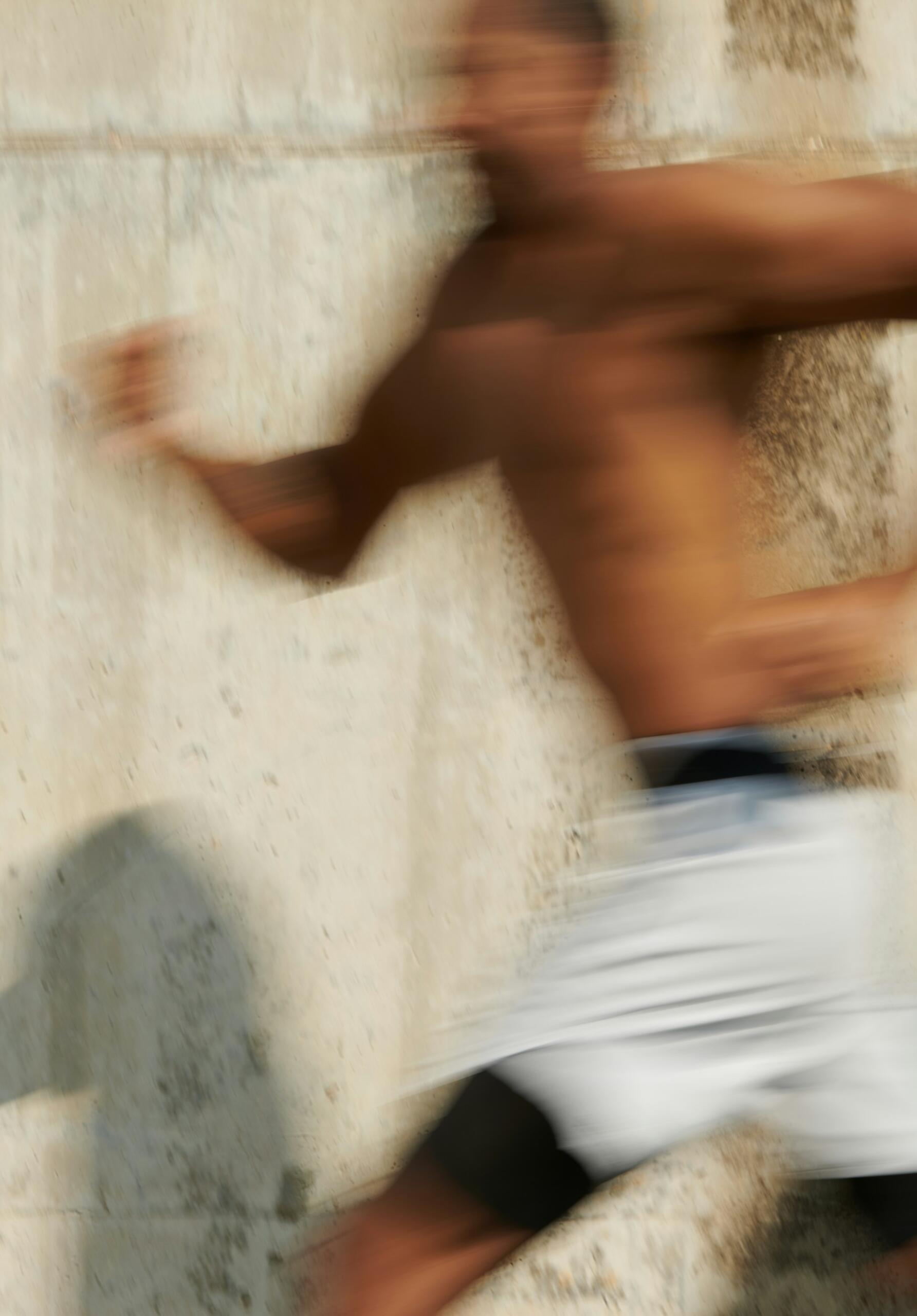 Imagen borrosa de un hombre sin camiseta y con pantalones cortos blancos corriendo junto a una pared con textura, que capta el movimiento y la velocidad.