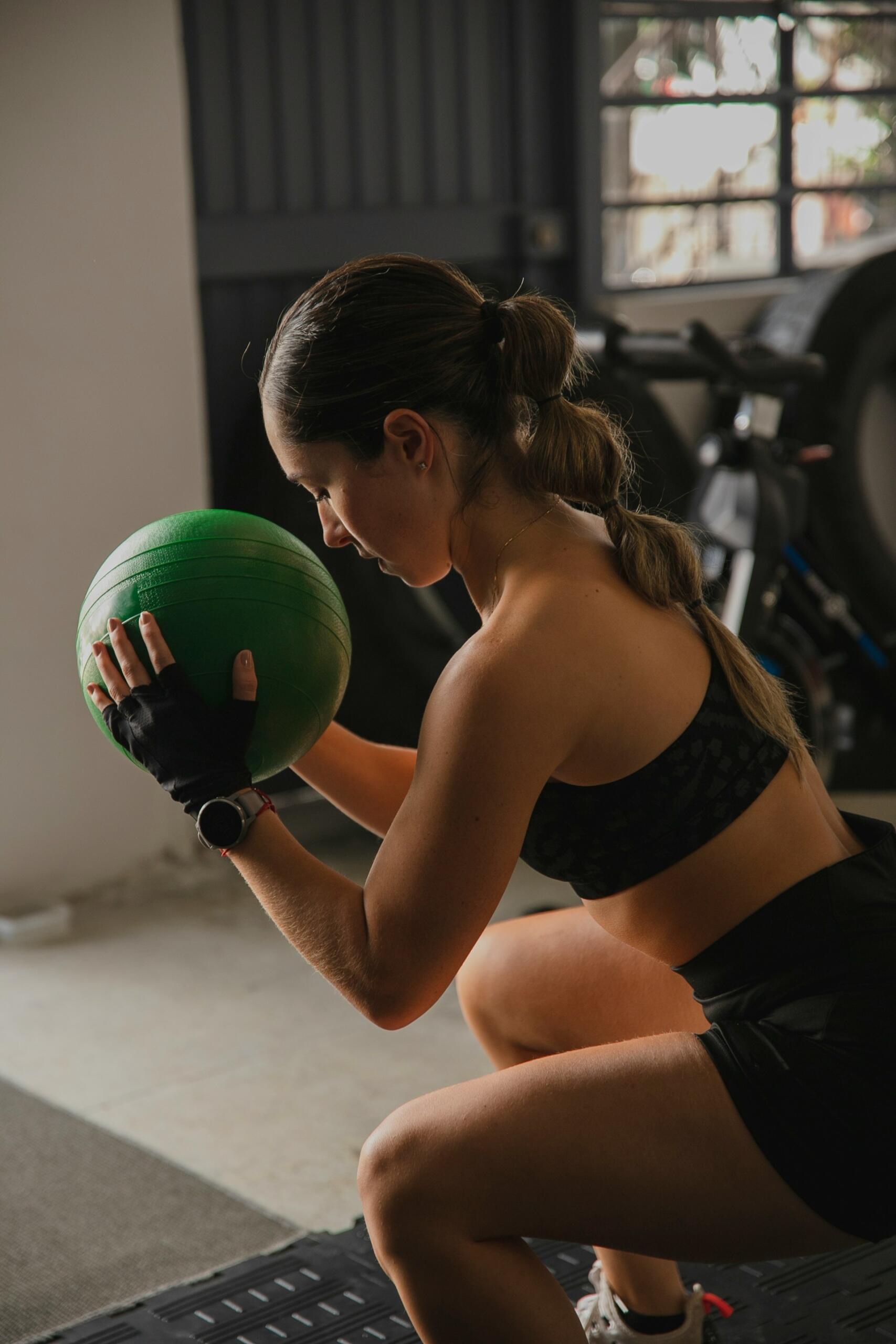 Una mujer vestida con ropa deportiva que sostiene un balón medicinal verde mientras hace sentadillas en un gimnasio.