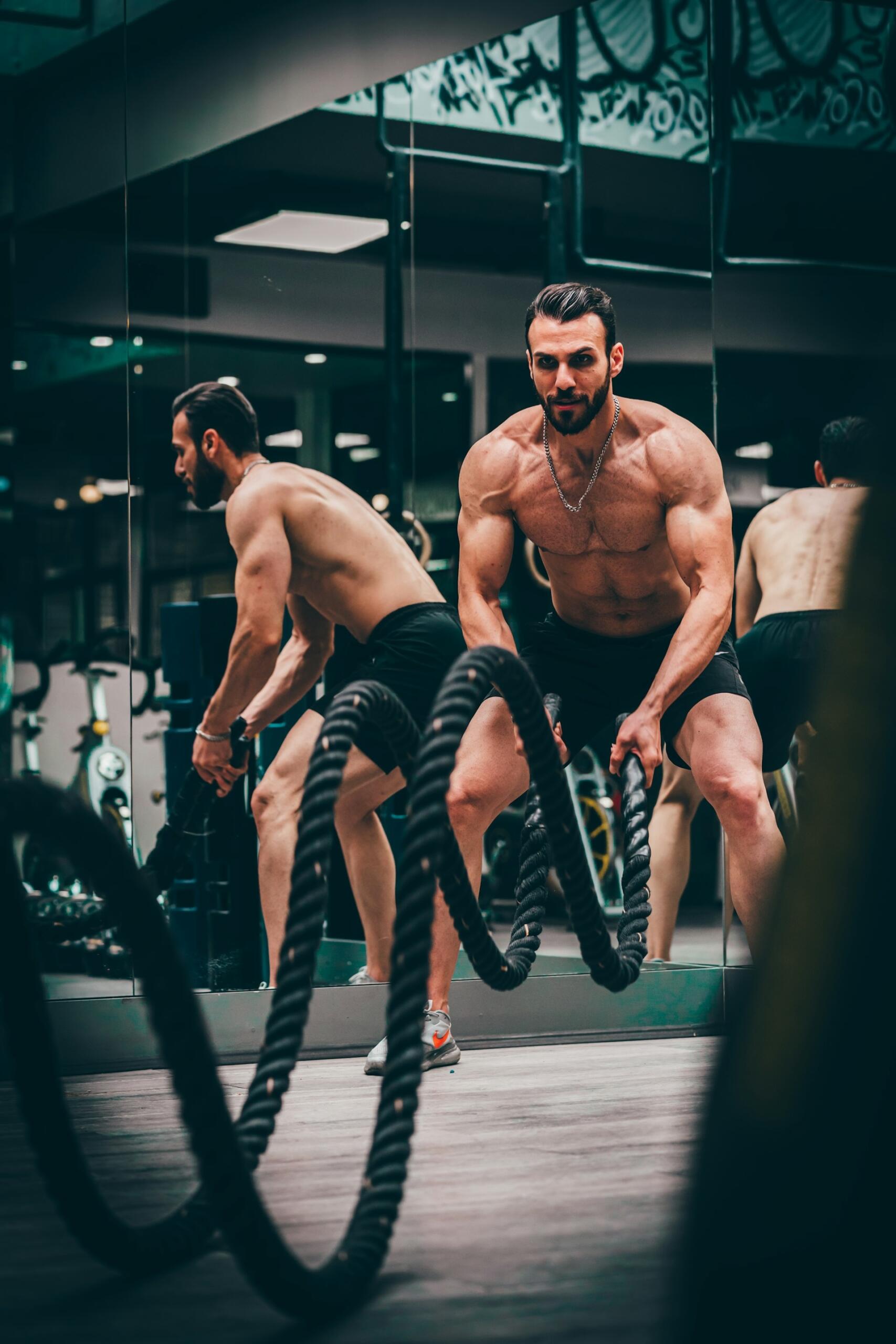 Un hombre musculoso y sin camiseta haciendo ejercicios con cuerdas de entrenamiento en un gimnasio con paredes de espejos, vestido con pantalones cortos negros y zapatillas blancas.