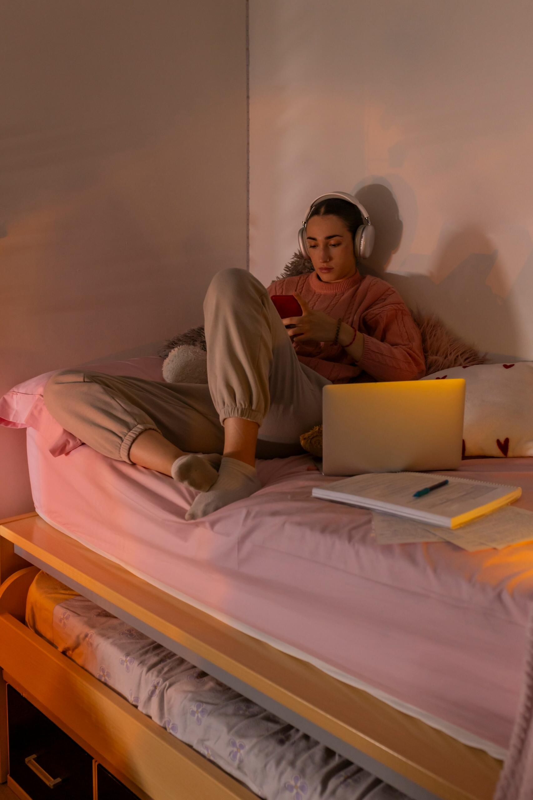 Persona con auriculares sentada en una cama con un ordenador portátil y un cuaderno, sosteniendo un smartphone.