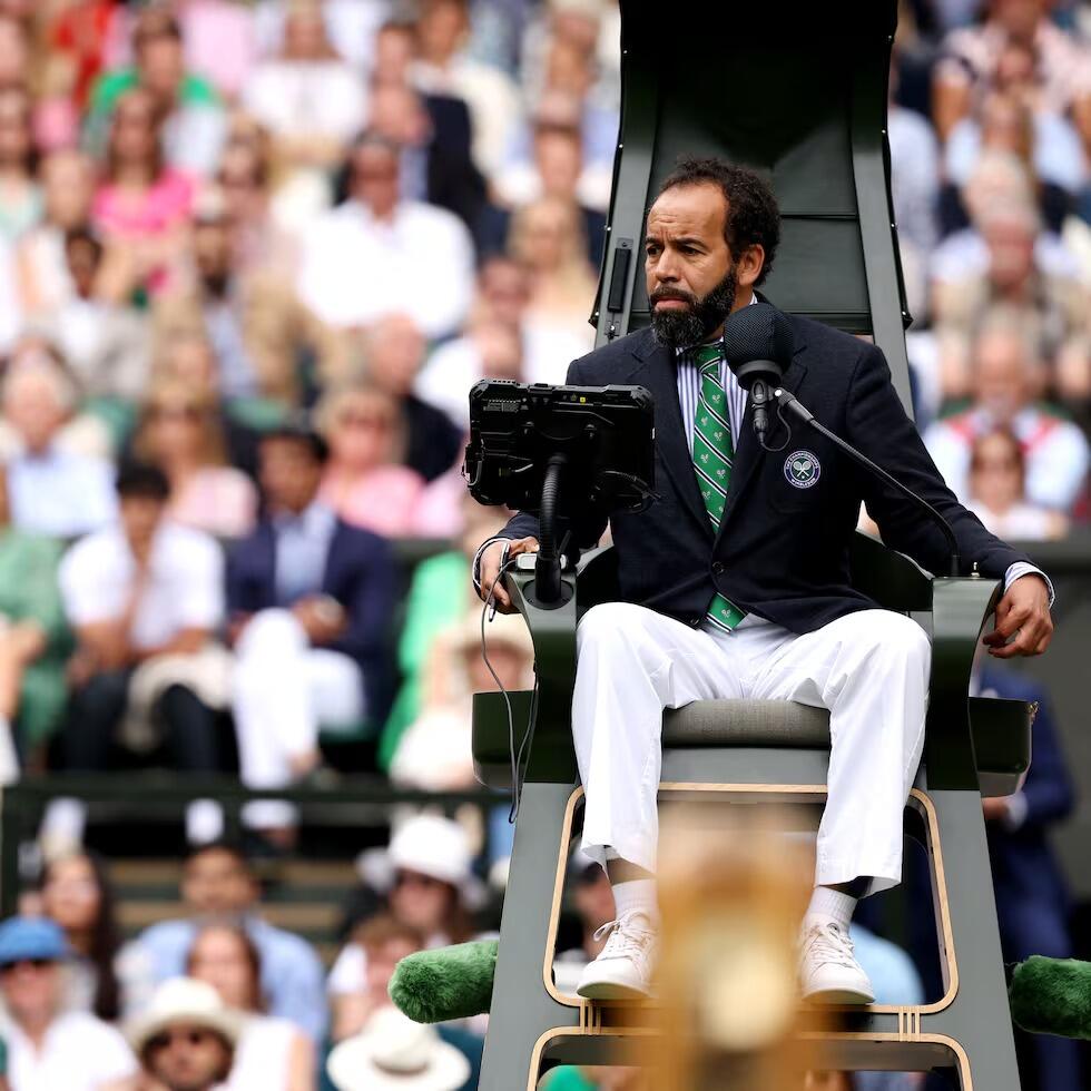 Un árbitro de tenis, vestido con una chaqueta oscura, corbata verde y pantalones blancos, está sentado en Wimbledon y se dirige a los jugadores mientras una multitud difusa observa la escena.