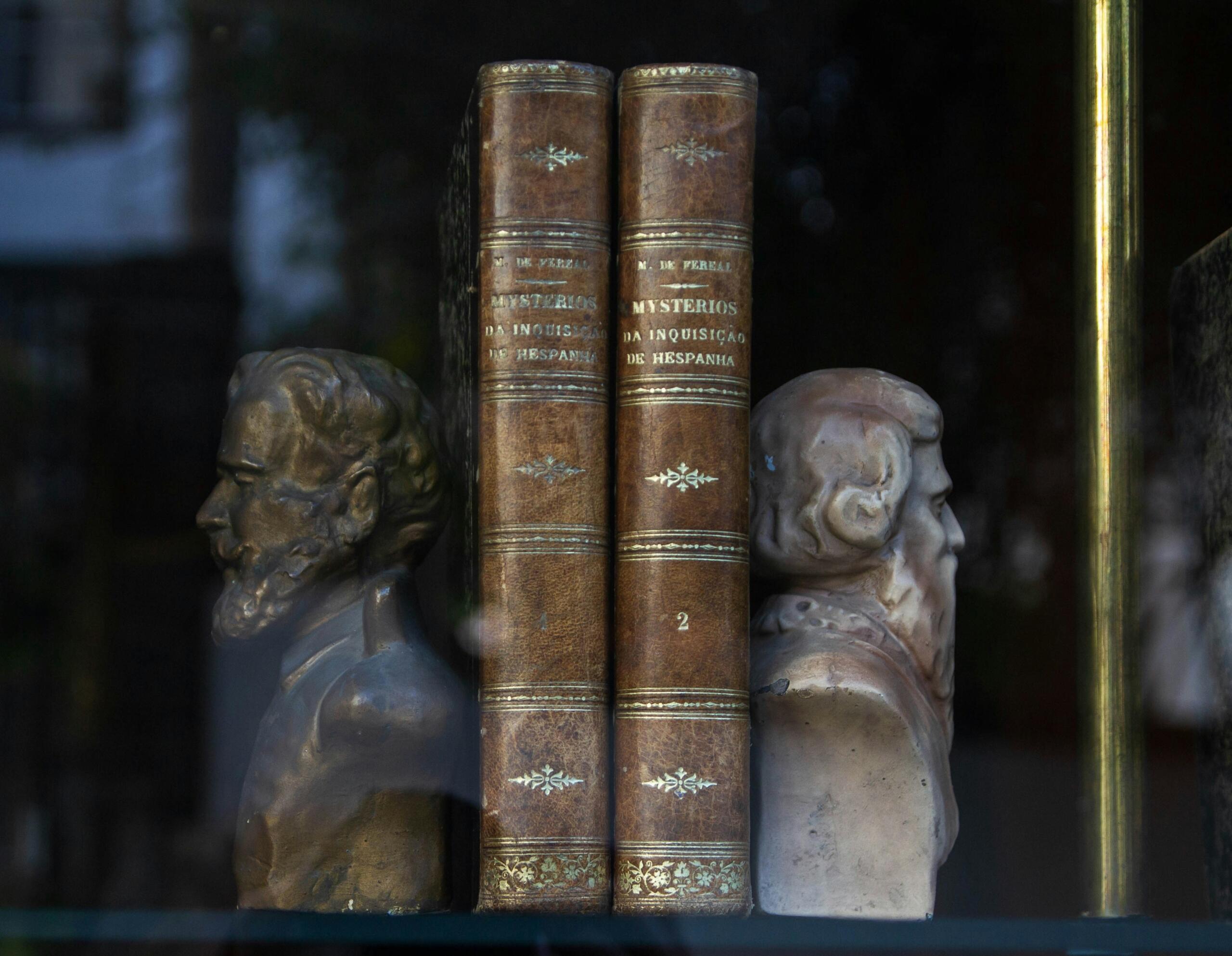 Dos libros antiguos titulados "Misterios da Inquisição de Hispania" junto a dos bustos de estilo clásico en un estante de exhibición.