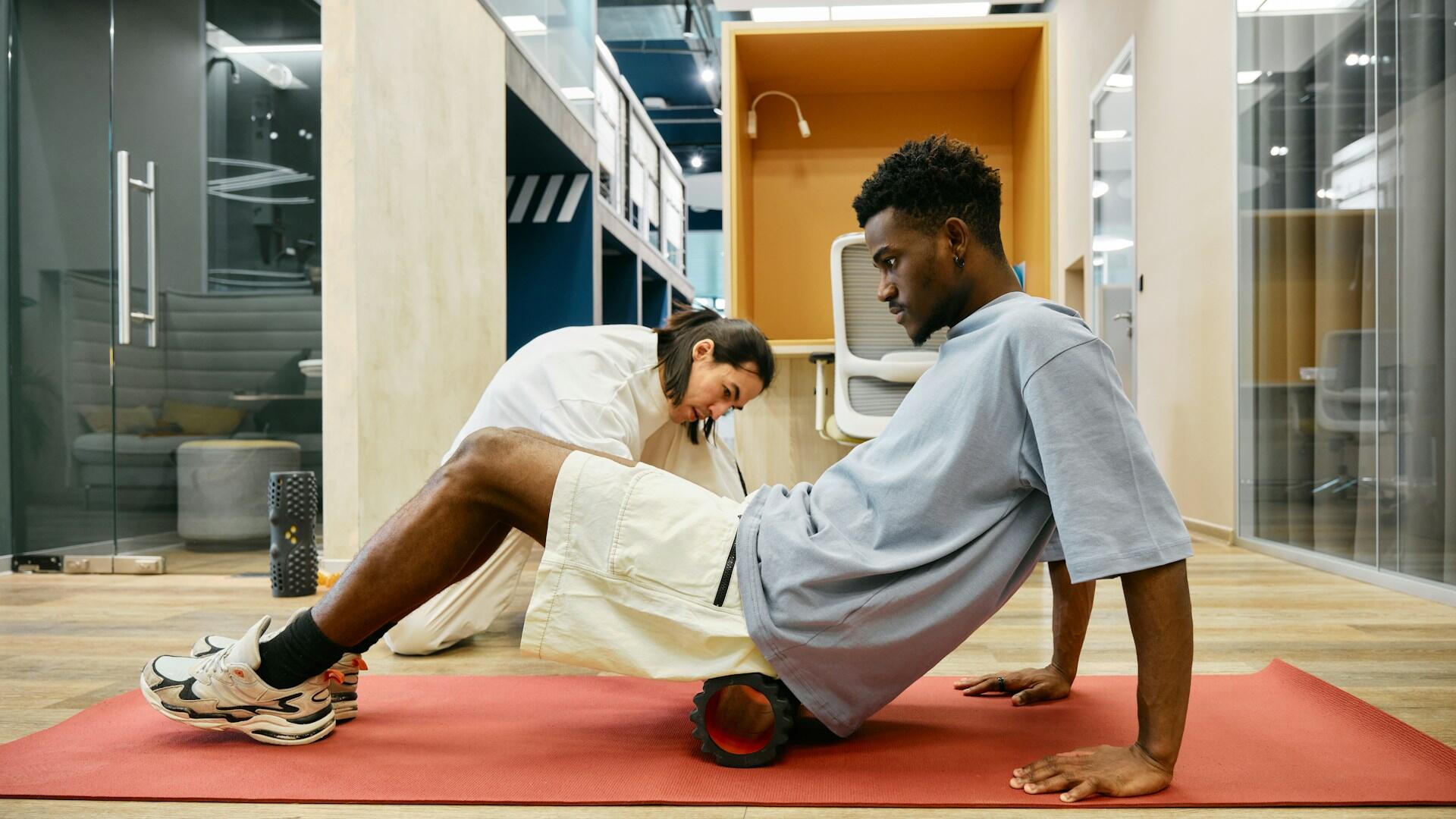 Una persona utiliza un rodillo de espuma para hacer ejercicio sobre una esterilla de yoga, mientras otra le ayuda en un espacio de trabajo moderno y bien iluminado.
