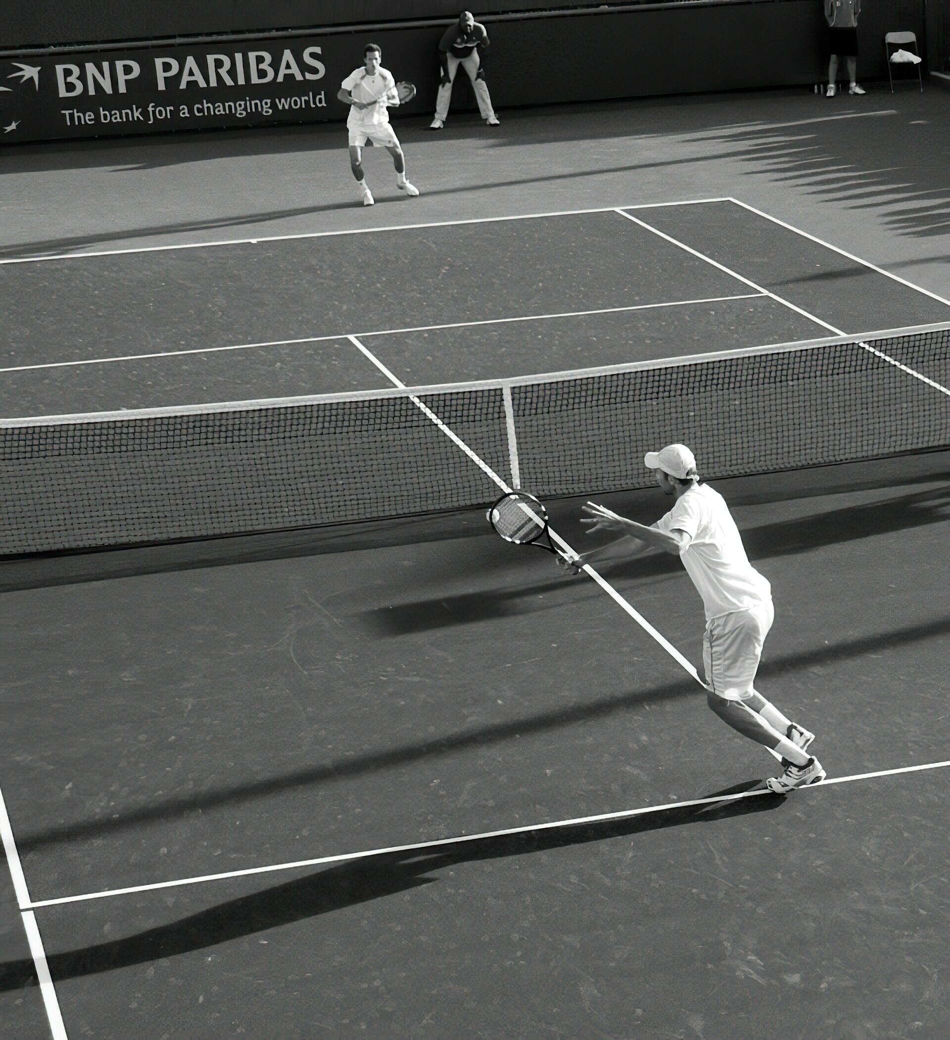 Partido de tenis en blanco y negro: dos jugadores intercambian golpes por encima de la red en una pista bañada por el sol, con el letrero de BNP Paribas y unas largas sombras que crean un efecto de profundidad.