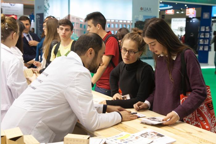 Un hombre con bata de laboratorio habla con dos mujeres en un concurrido stand de una feria. Los visitantes, en su mayoría jóvenes, observan con atención la exposición, creando una escena animada.