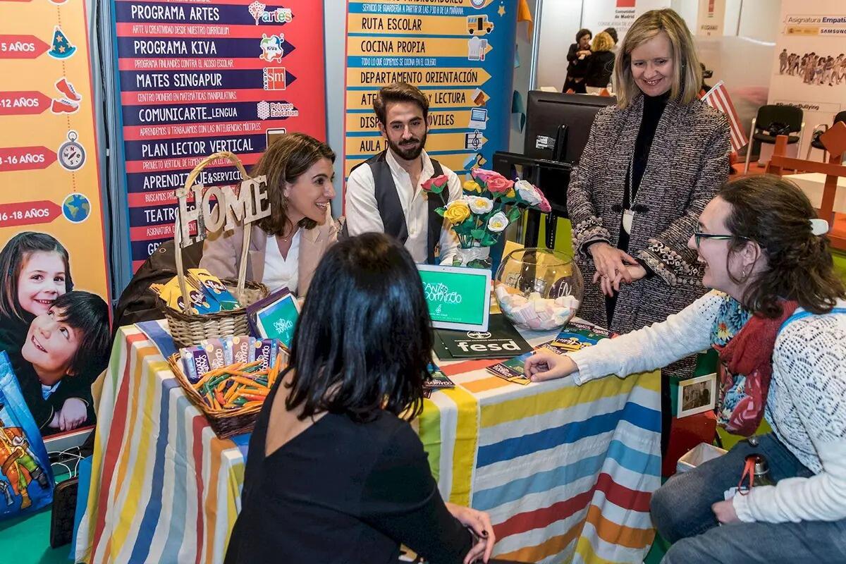 Un grupo de personas interactúa alrededor de un colorido stand en una feria educativa. La mesa, cubierta con un mantel a rayas, muestra materiales educativos. Las caras sonrientes transmiten un ambiente acogedor y atractivo.