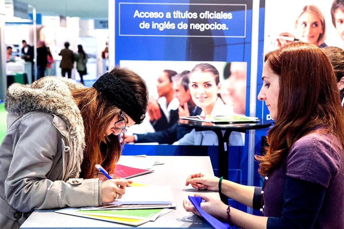 Una mujer con un abrigo de invierno y una cinta en la cabeza se inclina sobre una mesa y escribe en unos papeles. Otra mujer con un jersey morado la ayuda. Detrás de ellas, hay un cartel que dice «Acceso a títulos oficiales de inglés de negocios» con una imagen borrosa de profesionales del mundo empresarial. El escenario es un entorno informativo y profesional.
