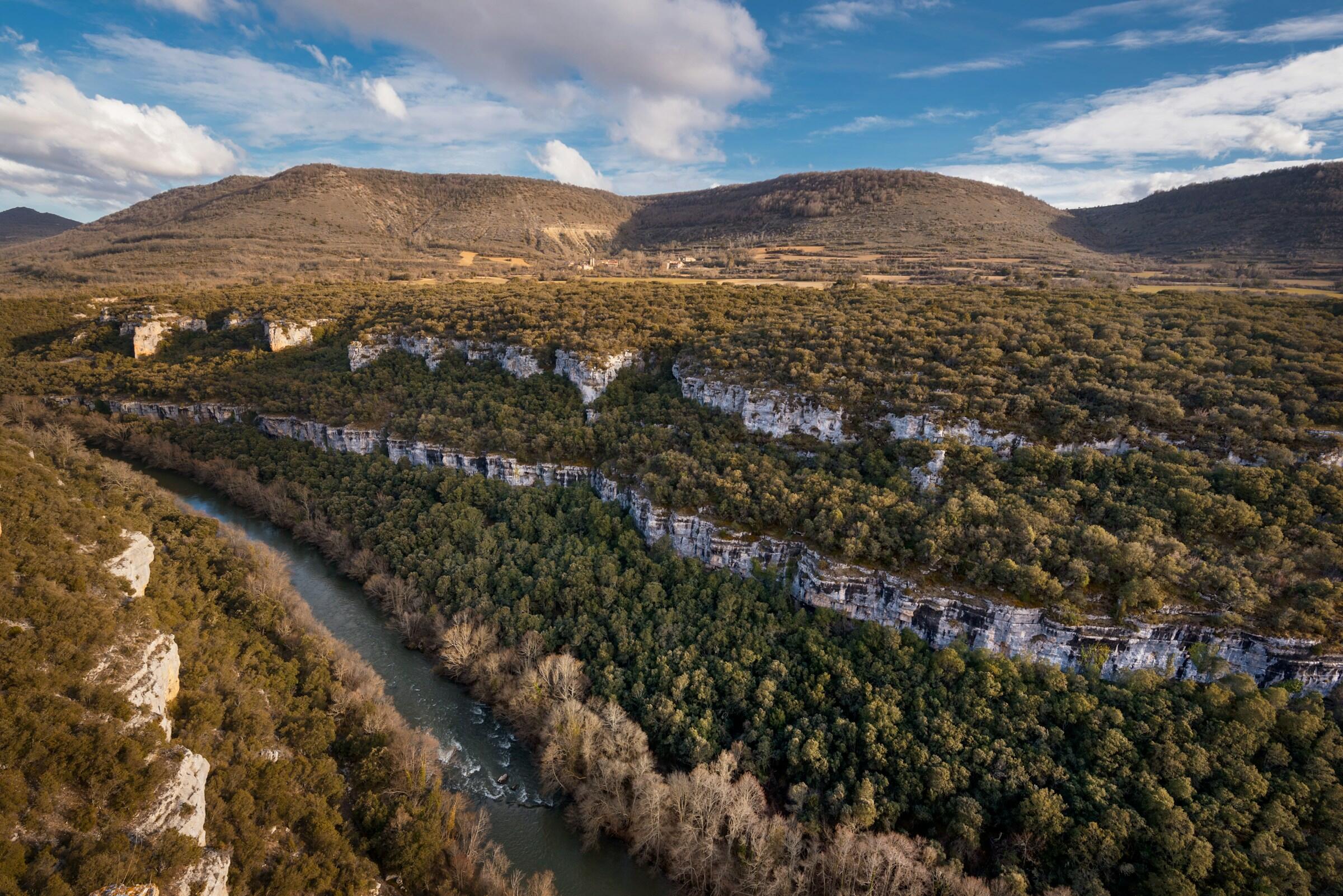 Vista aérea de un exuberante valle verde con el rio ebro serpenteante, rodeado de acantilados rocosos y colinas lejanas bajo un cielo parcialmente nublado.