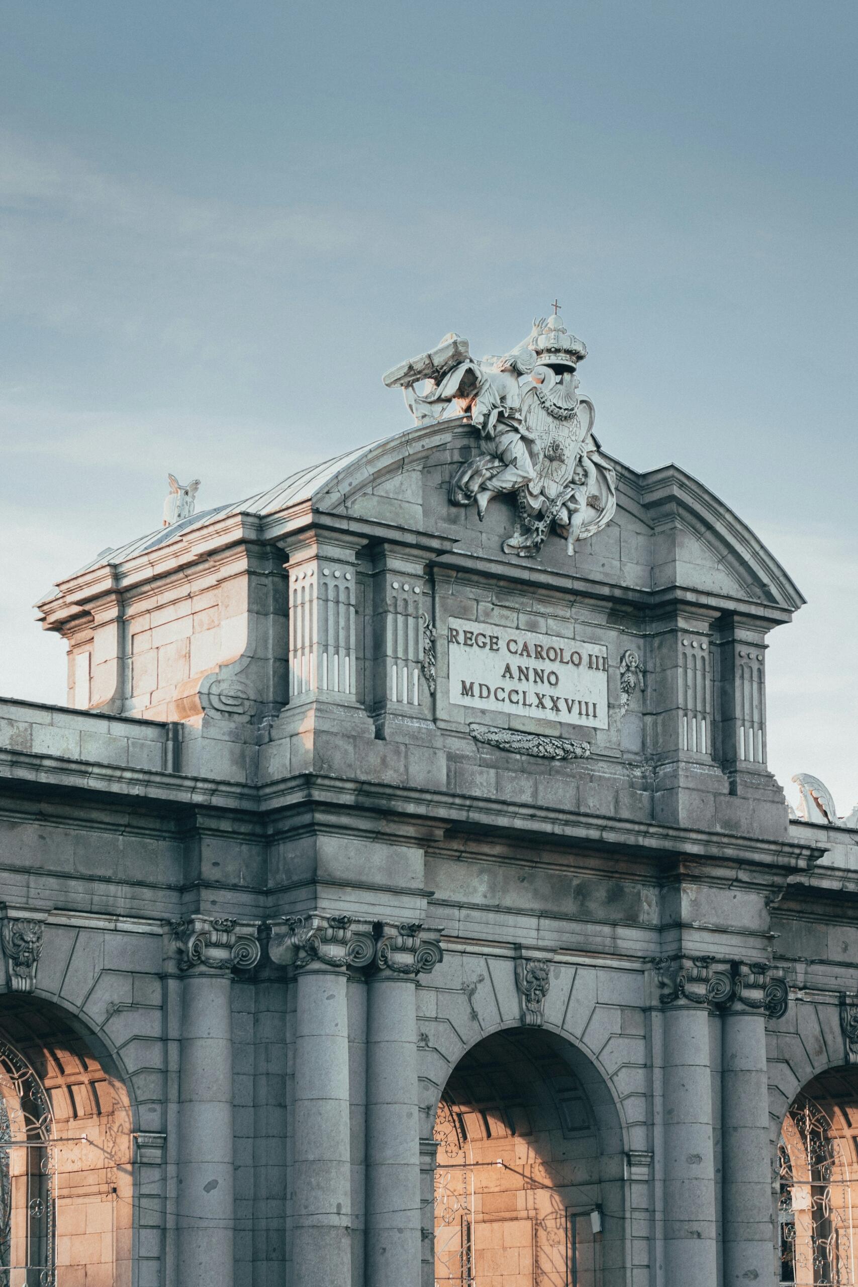 Detalle de un gran arco de piedra la puerta de alcala de madrid con tallas ornamentales y una placa con la inscripción «Rege Carolo III Anno MDCCLXXVIII» sobre un cielo azul.