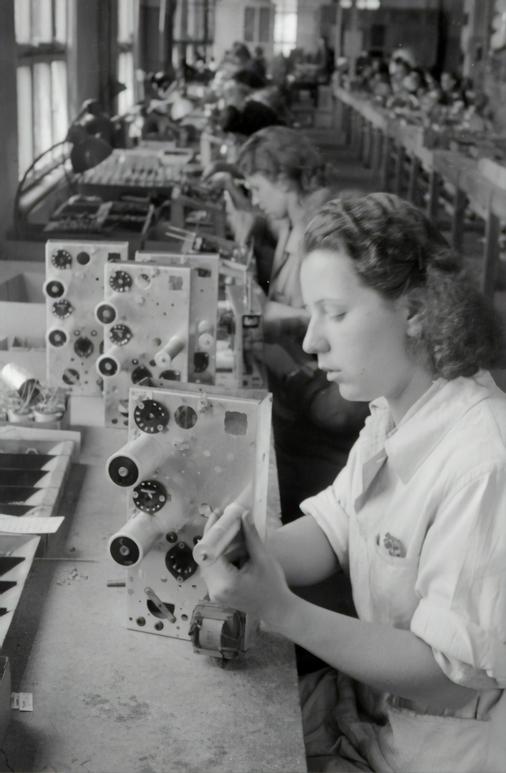Unas mujeres ensamblan equipos electrónicos en un largo banco de trabajo, centrándose en los intrincados componentes en el histórico entorno de una fábrica.