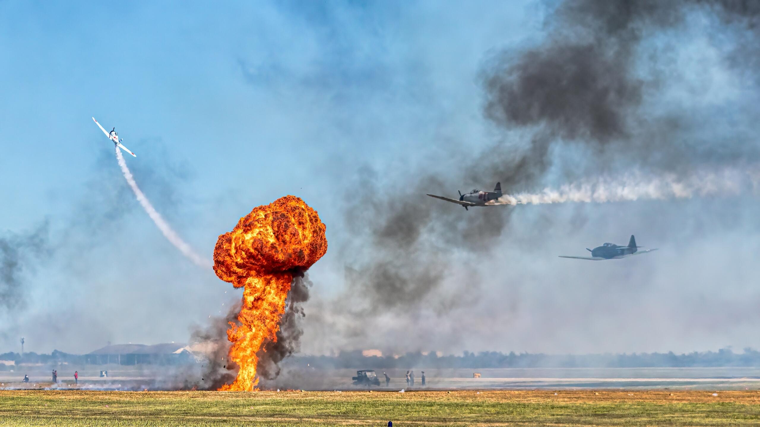 Una enorme explosión estalla en tierra mientras tres aviones realizan maniobras aéreas por encima, creando estelas de humo contra un cielo despejado.