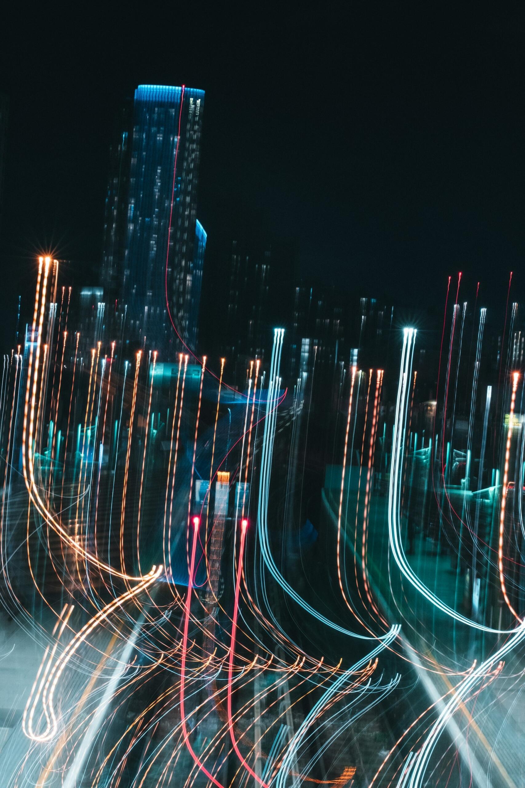 Paisaje urbano nocturno abstracto con rastros de luz de diversos colores, que crean un efecto dinámico de neón. En el fondo se divisa vagamente un rascacielos.